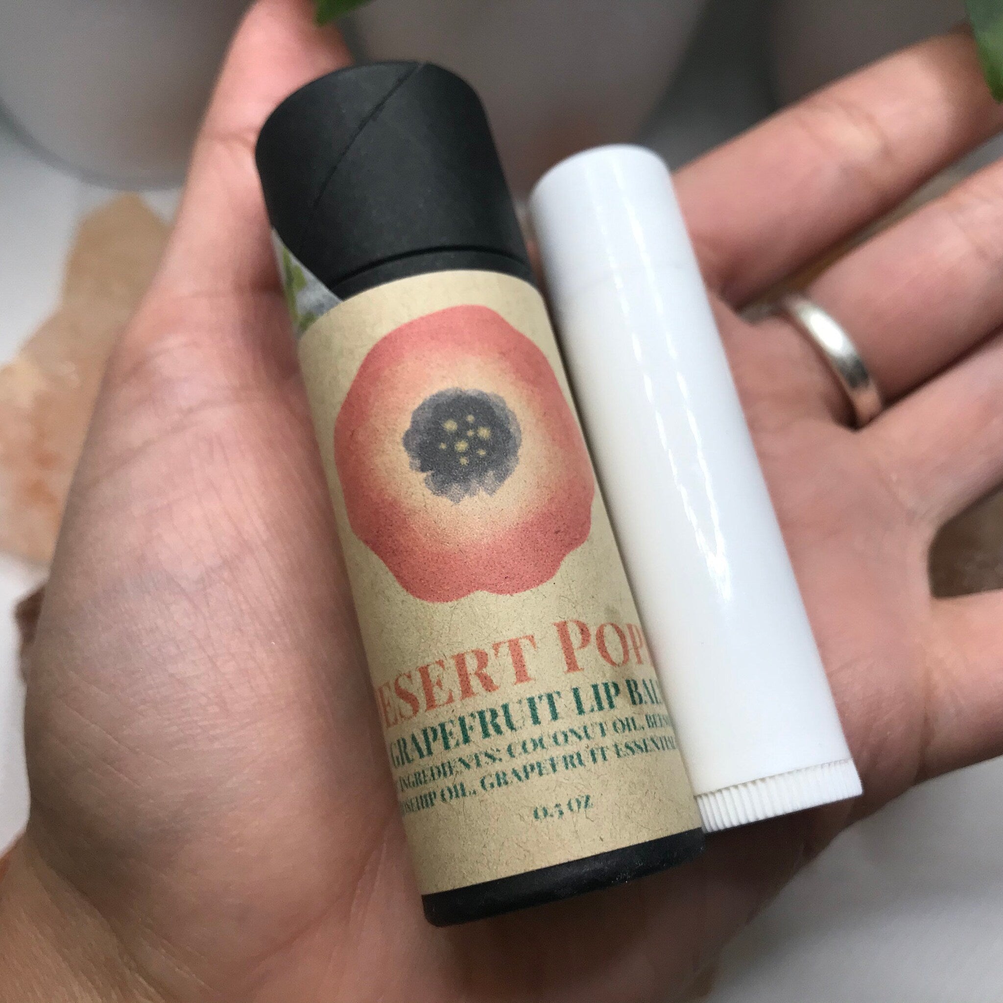 Lip balm tubes in hand, one with floral design on brown paper, one white, cylindrical, and open