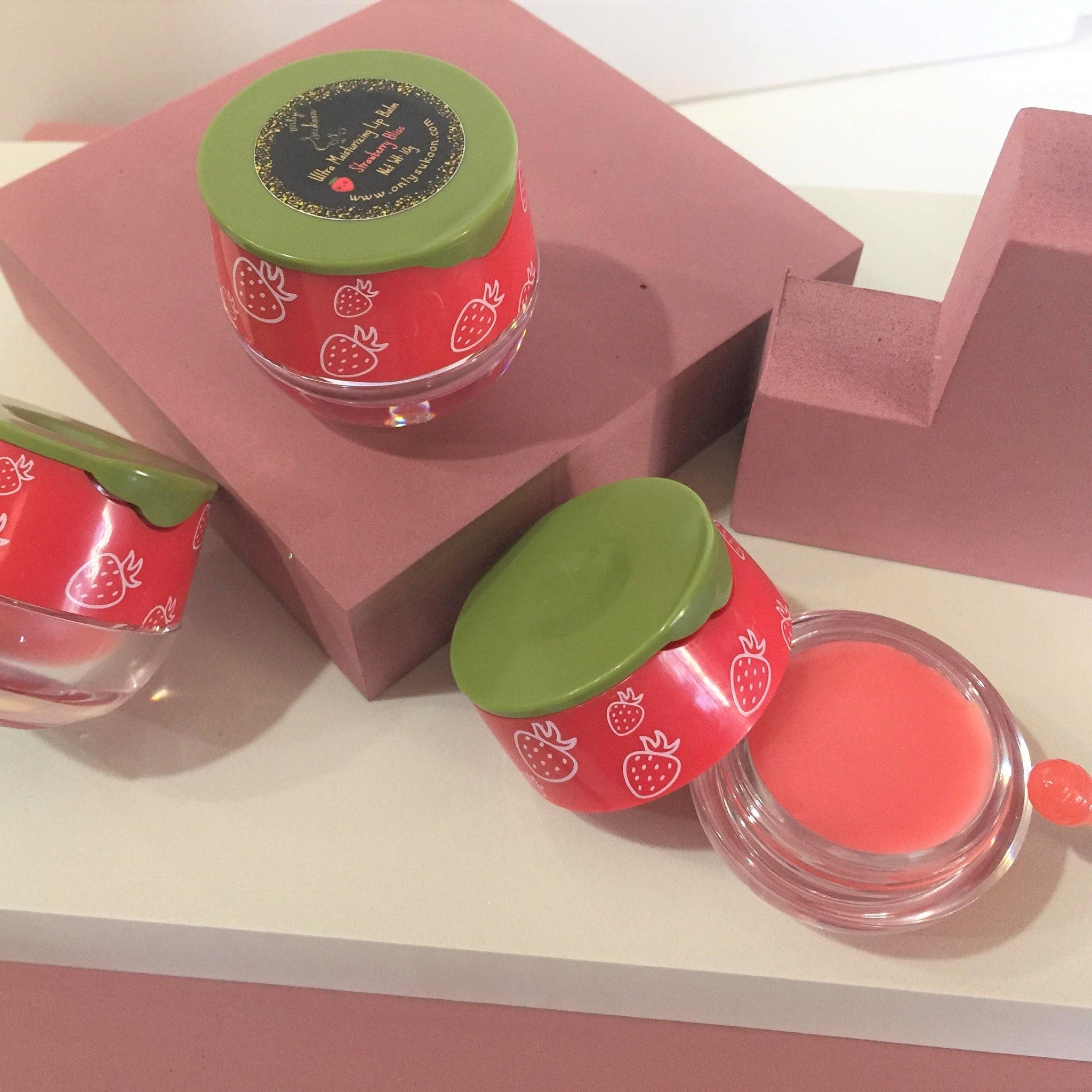 Lip balm with red strawberry pattern, green lid, and clear container, Strawberry Bliss label visible