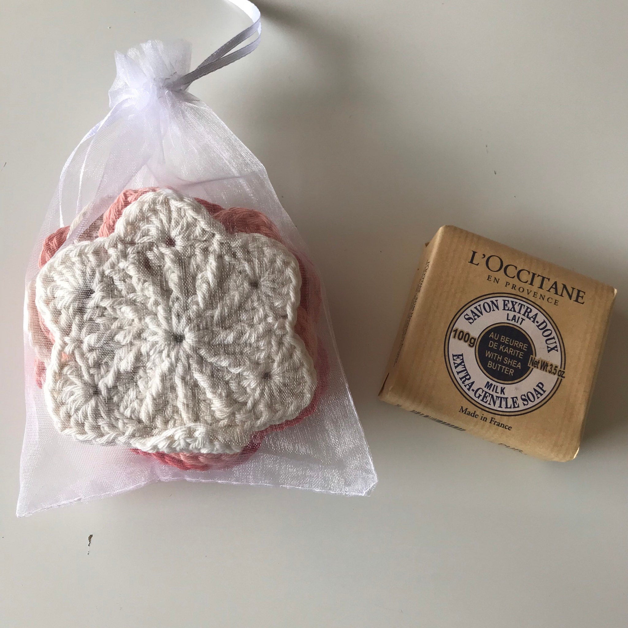 Crochet face scrubbies with white, pink, and cream colors in a sheer bag, next to L'Occitane soap bar