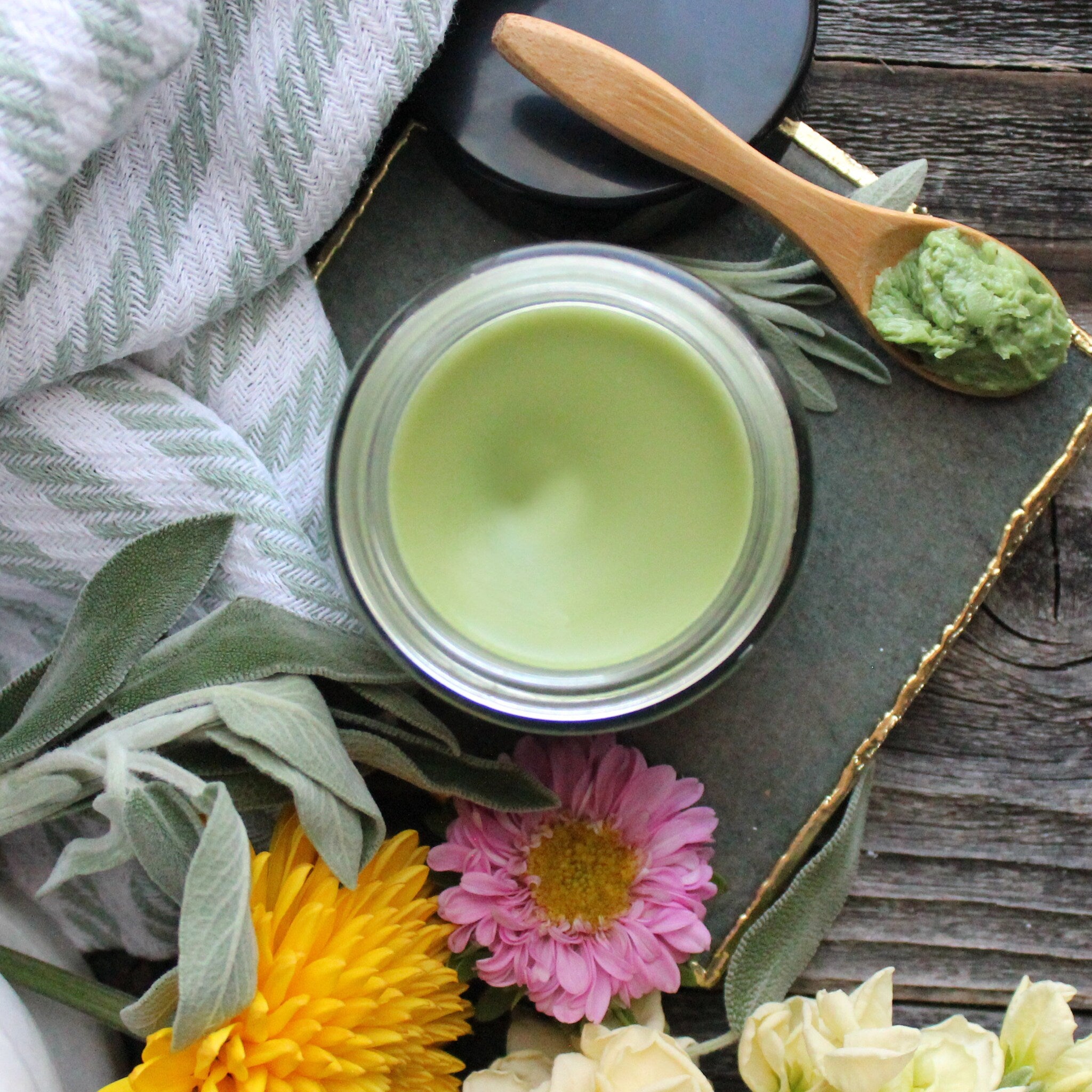 Green balm in glass jar with wooden spoon and flowers, on wooden surface, with patterned cloth