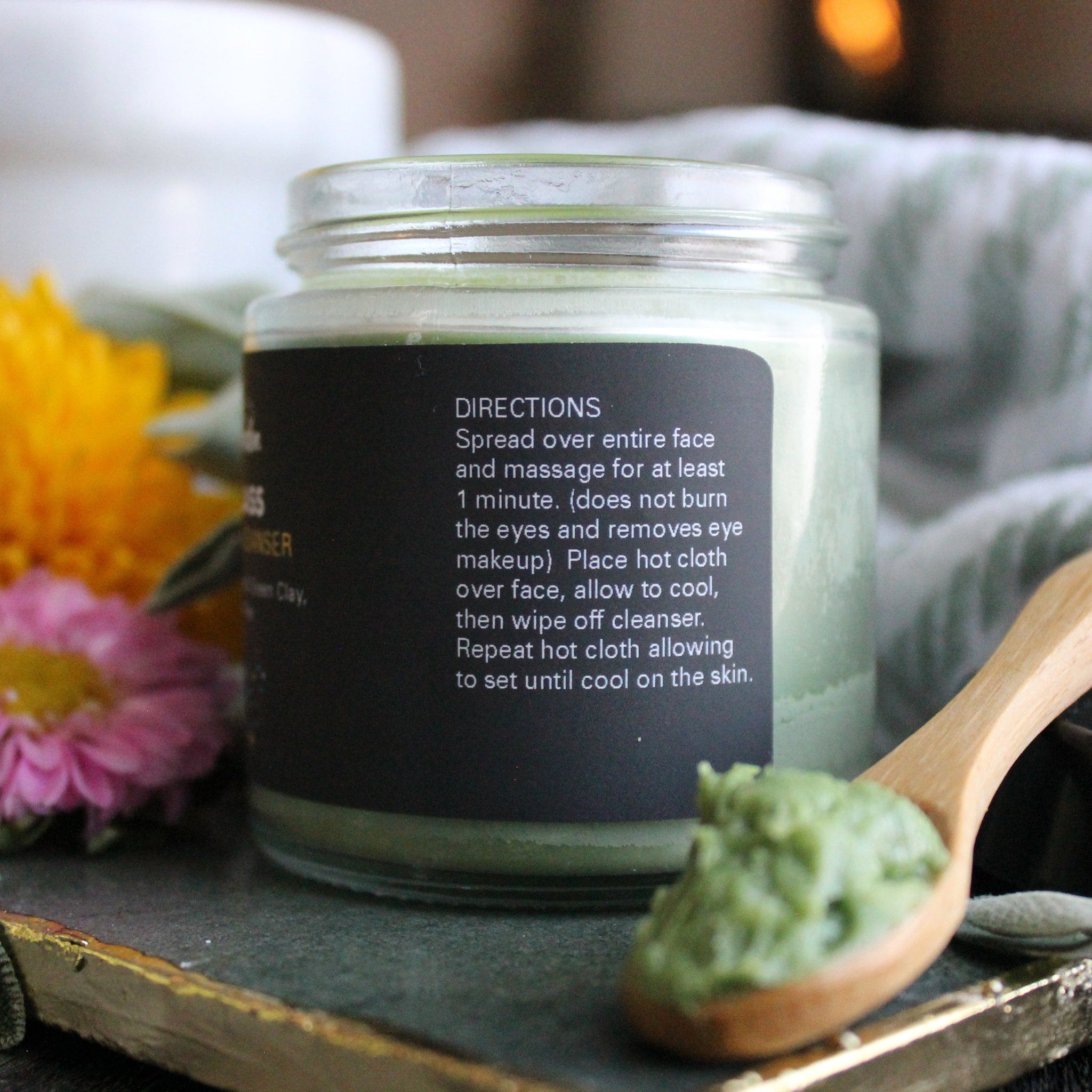 Green cleanser in glass jar with black label, wooden spoon with green product, and flowers