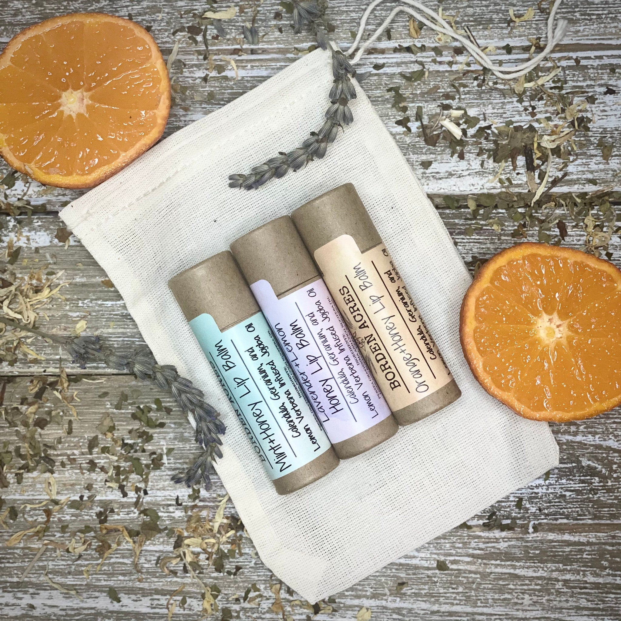 Lip balms in brown tubes with colorful labels, on white cloth with orange slices and herbs