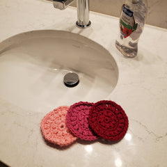 Face Scrubbies, three crochet rounds in pink, fuchsia, and red, near a sink