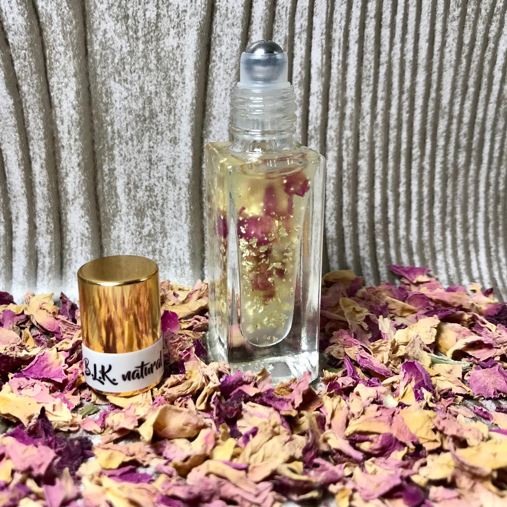 Roll-on perfume bottle with clear glass, gold cap, and B.L.K. natural label, surrounded by dried rose petals