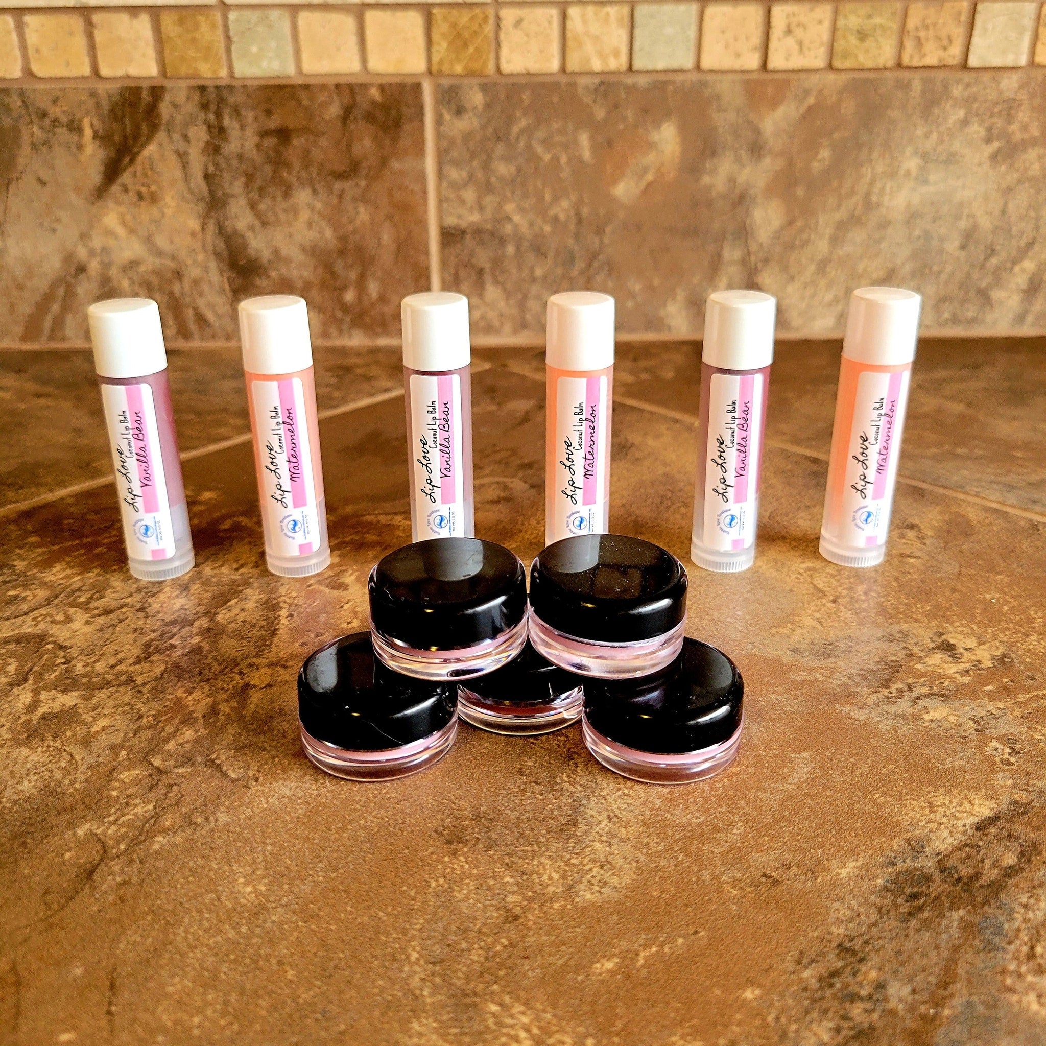 Lip balm tubes with white caps, pink labels, and black jars on a brown surface