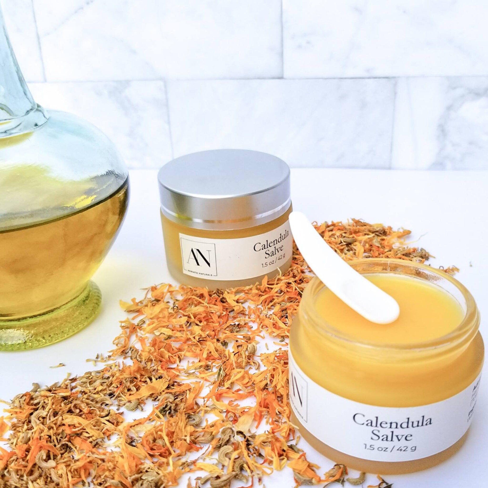 Calendula salve jars with silver lids, yellow cream, and orange flower petals on white surface