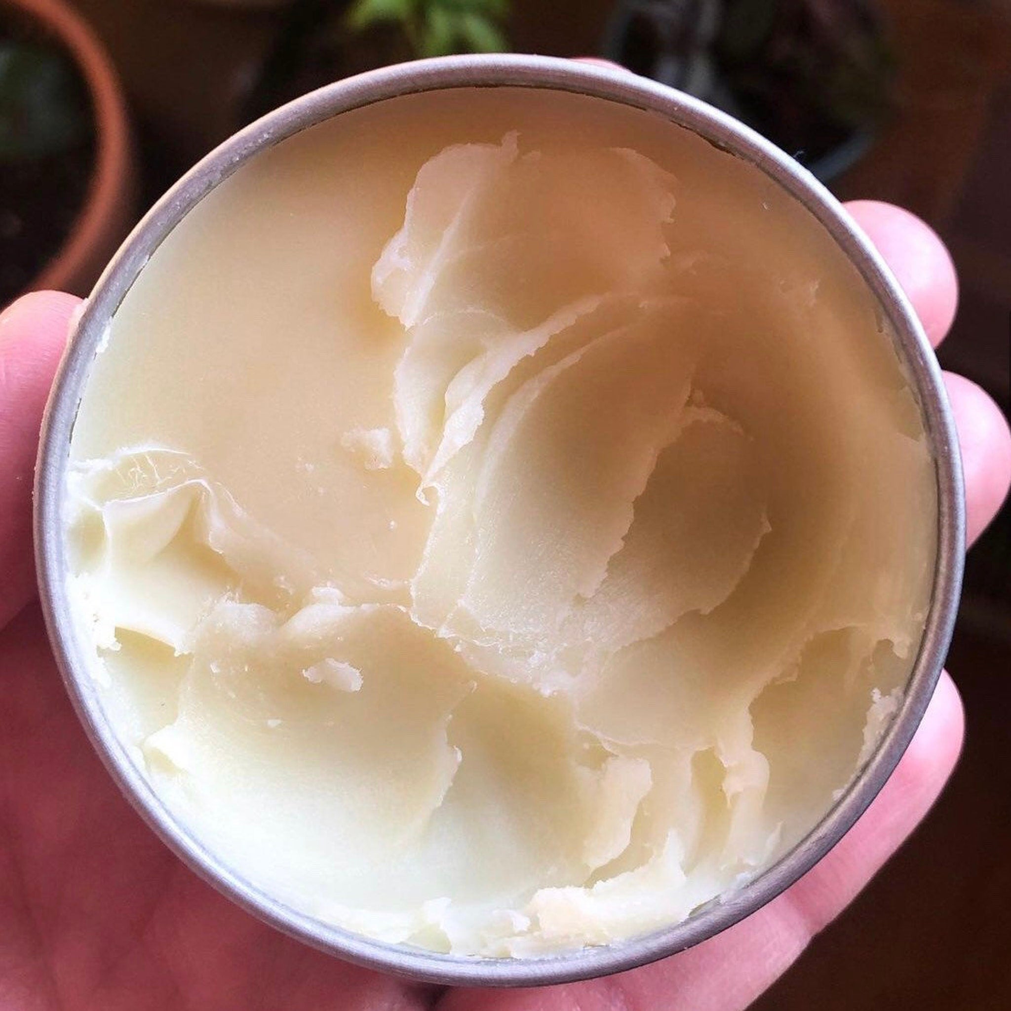 Creamy white balm in a silver metal tin, held in hand, showing texture and color.