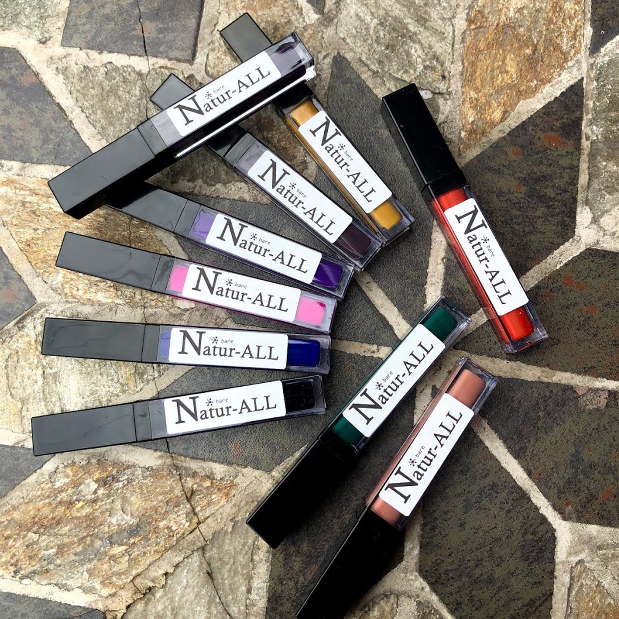 Lip gloss tubes in various colors with black caps and Natur-ALL label, arranged on stone surface