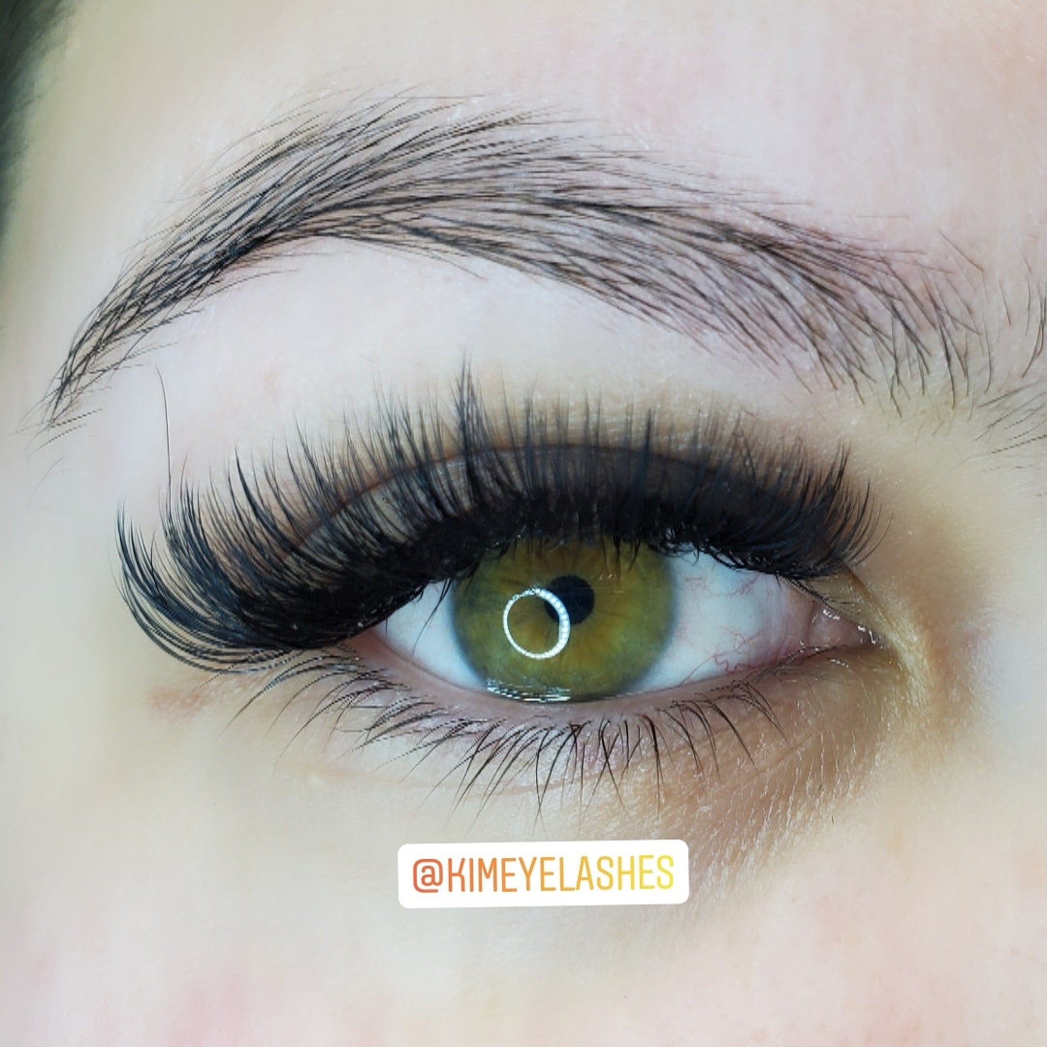 Eyelash extensions with long black lashes, green eye, and dark eyebrow, @KIMEYELASHES logo visible