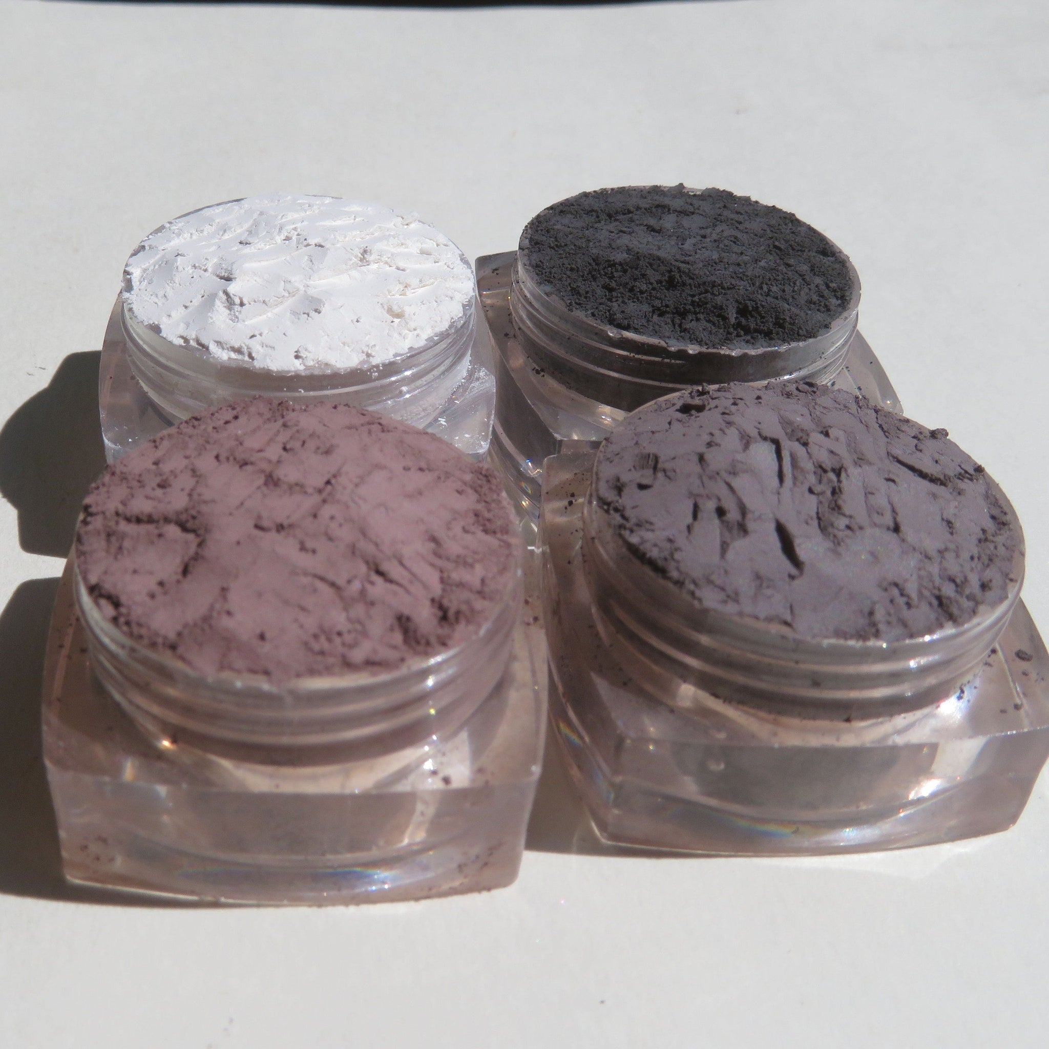 Close-up of four loose mineral eyeshadows in glass jars showing matte and shimmer textures in earthy tones.