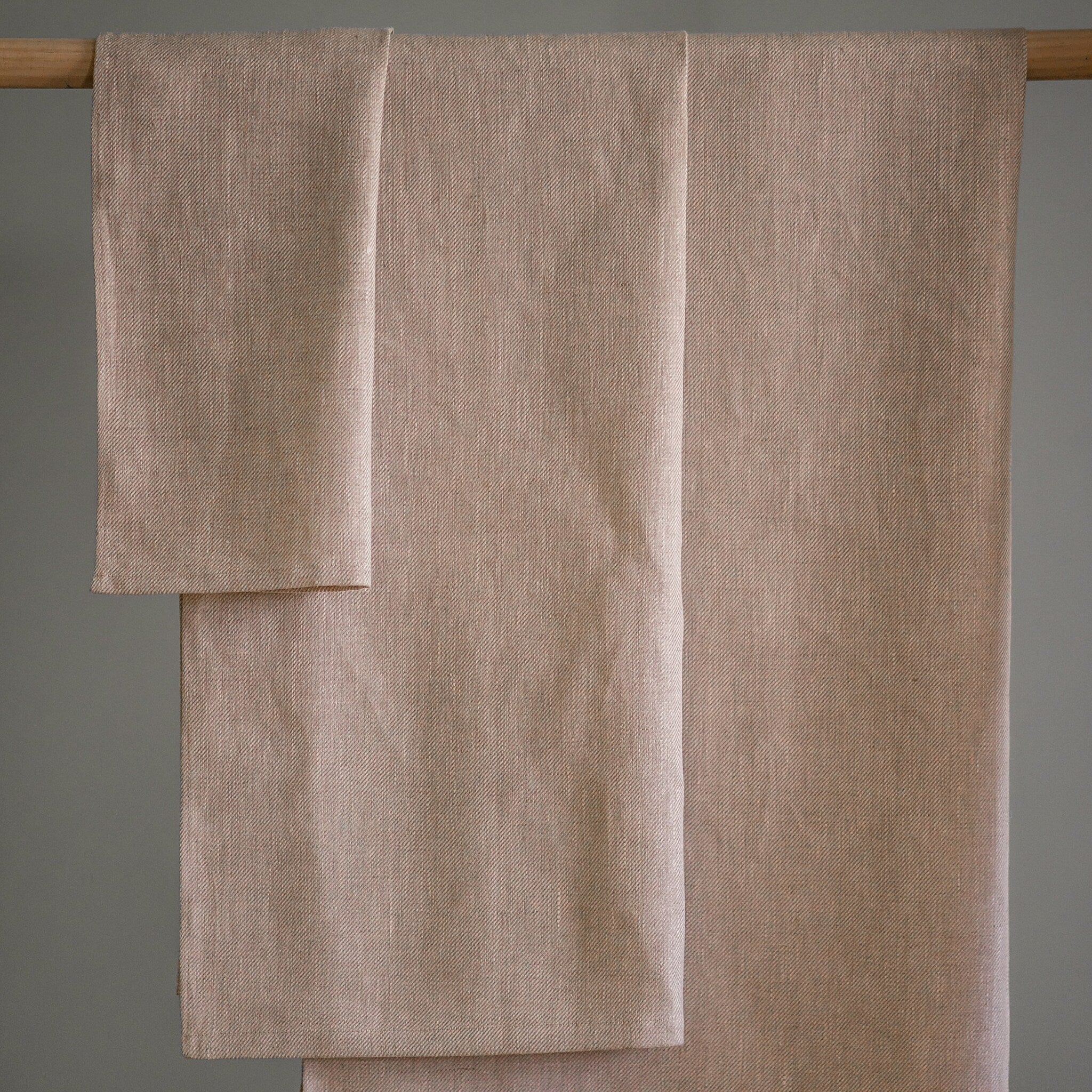 Beige linen fabric with diagonal weave, hanging from wooden rod, neutral background
