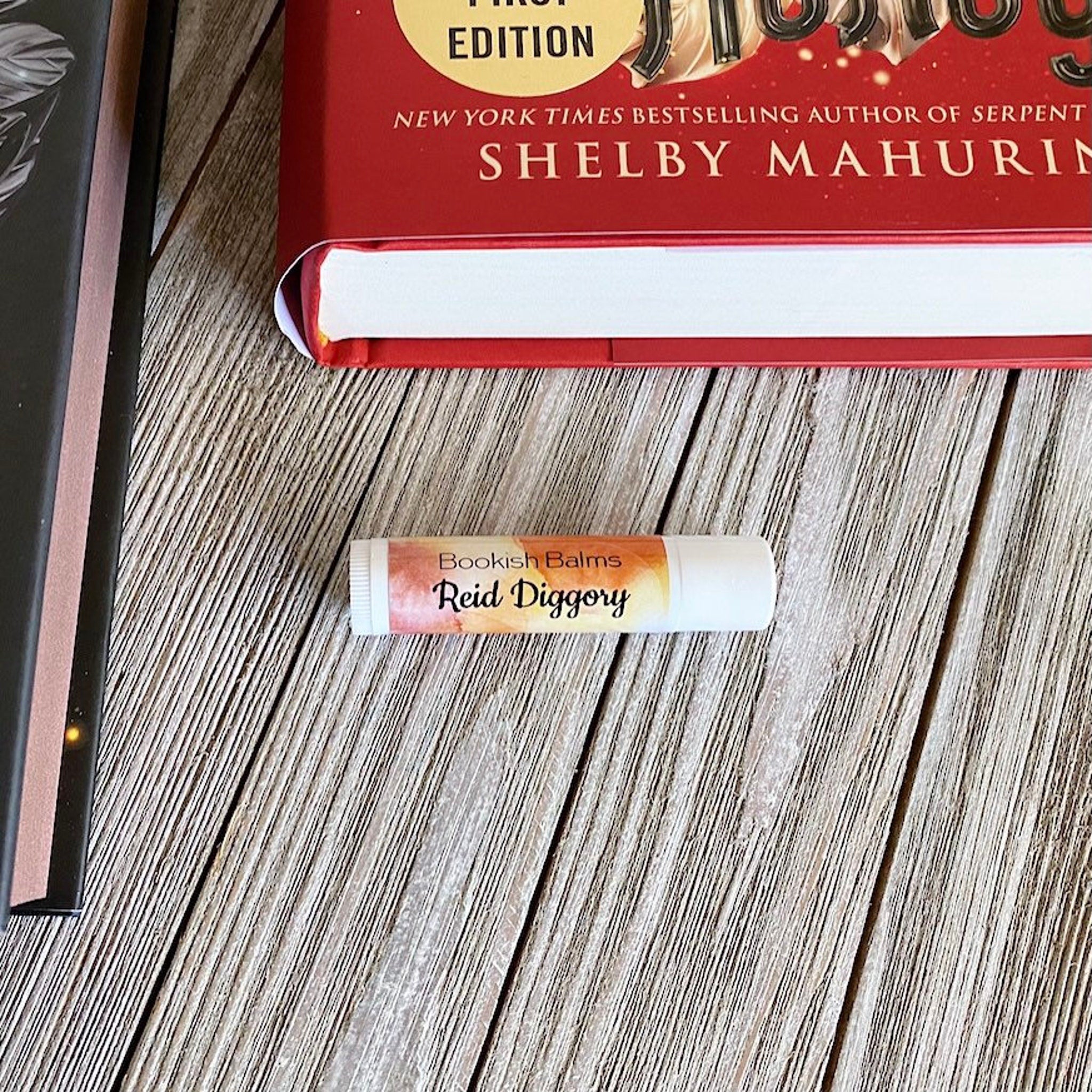 Lip balm tube with white, orange, and Bookish Balms text on wooden surface