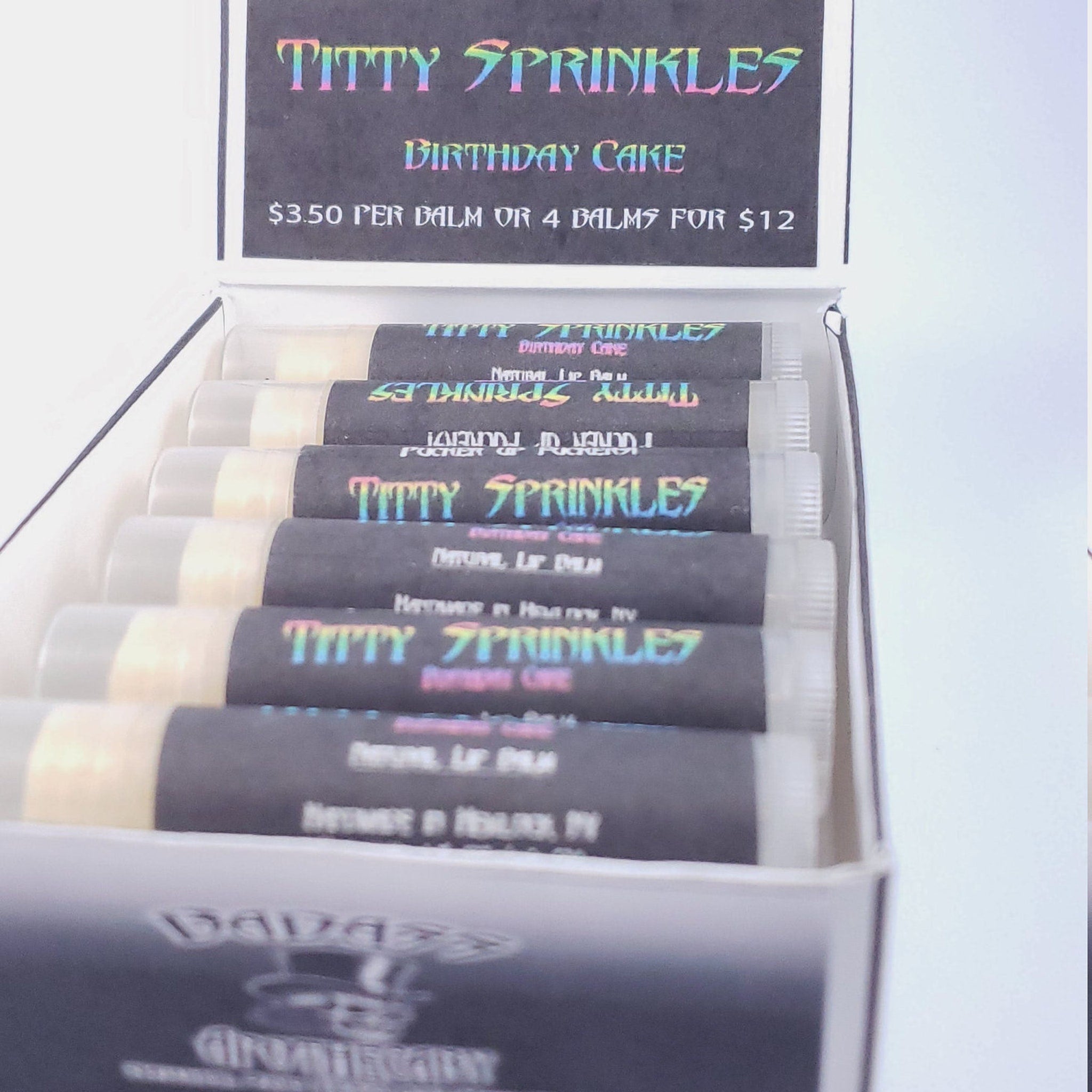 Lip balm tubes with black labels, clear plastic, and "TITTY SPRINKLES" text in rainbow colors, inside a white box