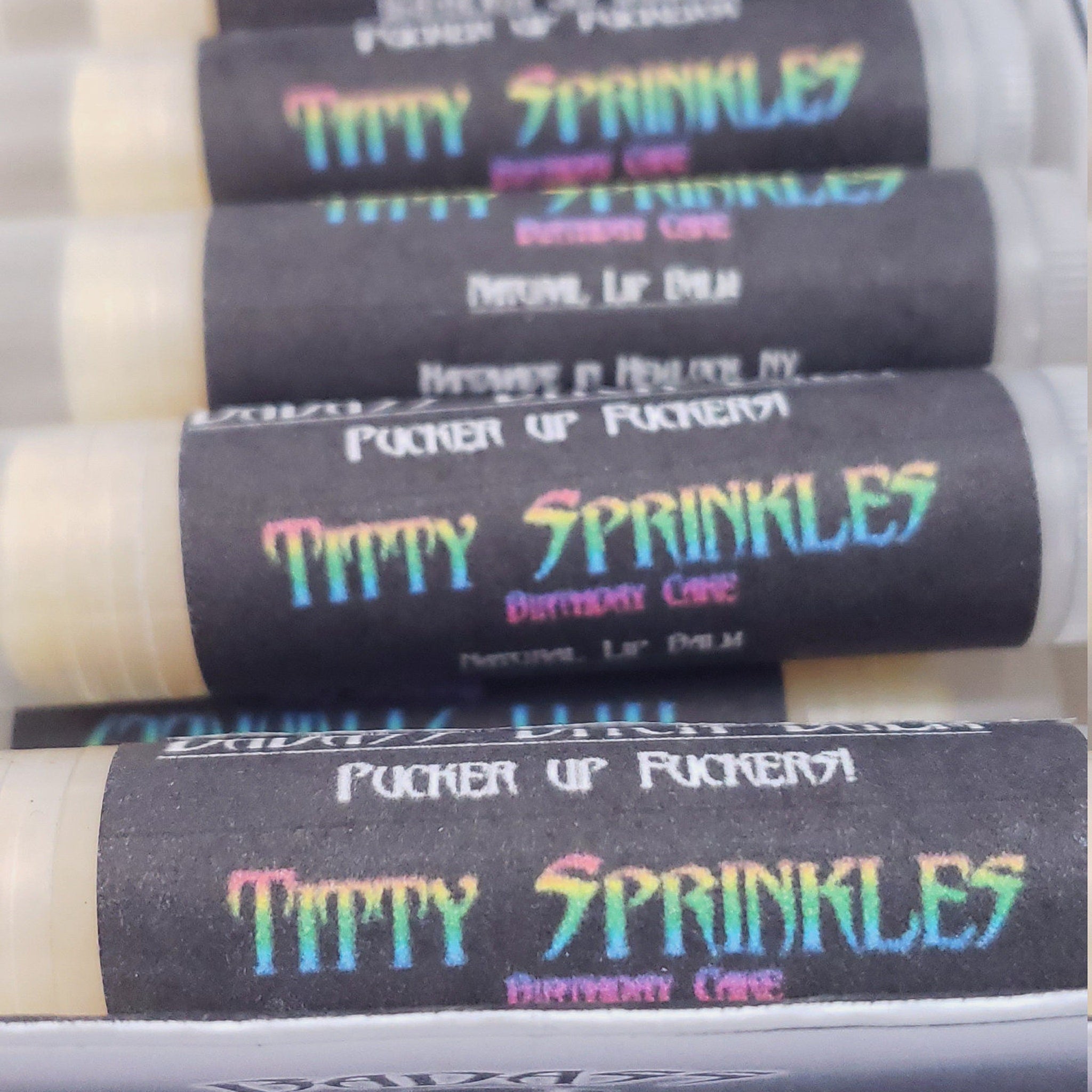 Lip balm sticks with black labels, rainbow text, and white packaging, stacked together
