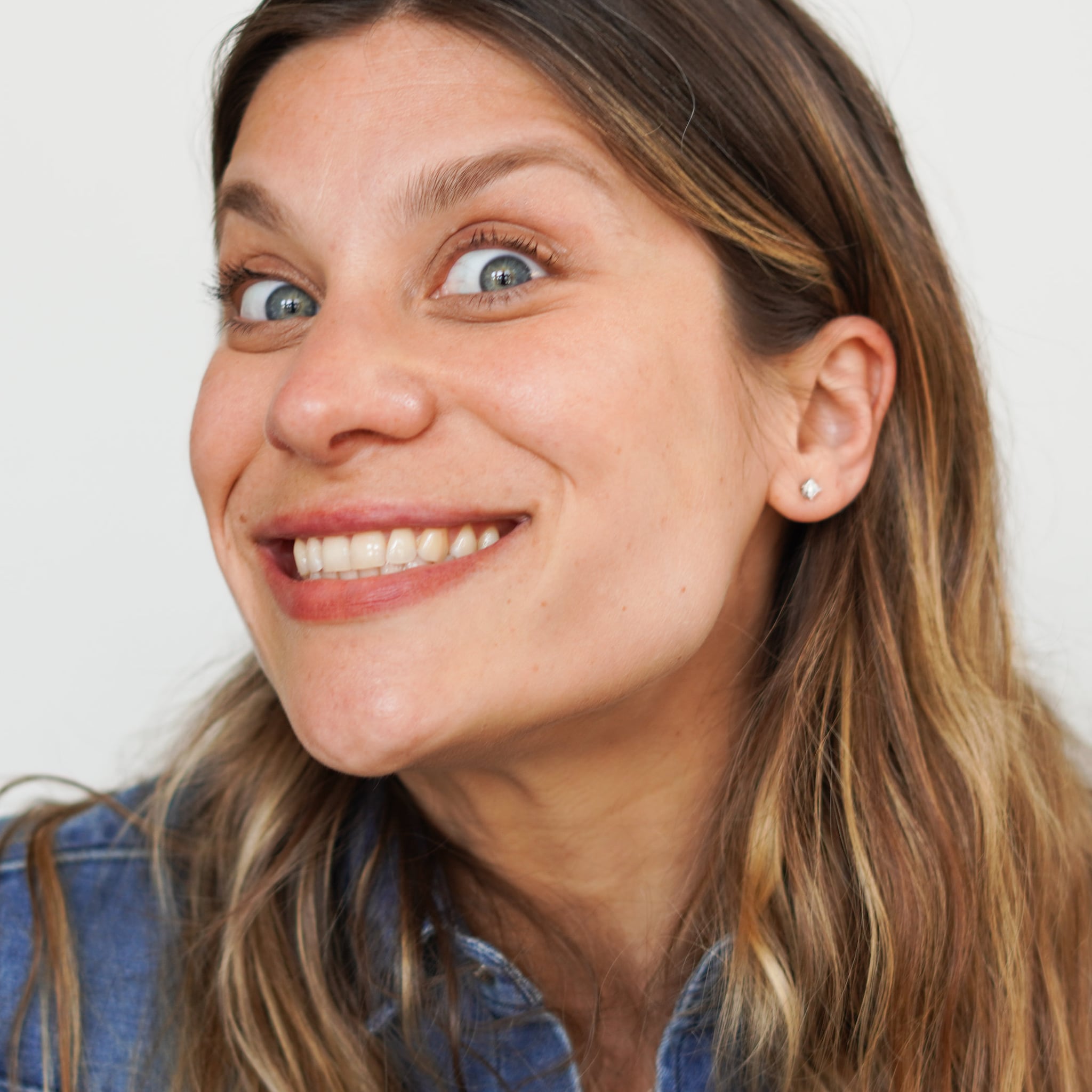 Woman with brown and blonde hair, wearing blue denim shirt, smiling with eyes wide open