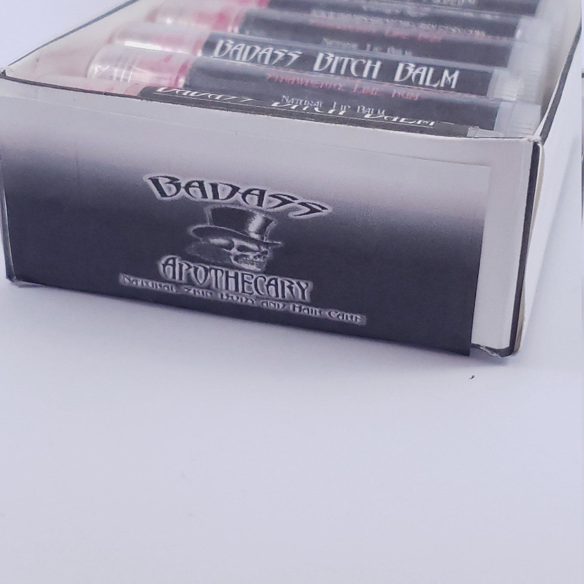 Lip balm tubes in clear plastic with black text, in a cardboard box with brand name "BADASS