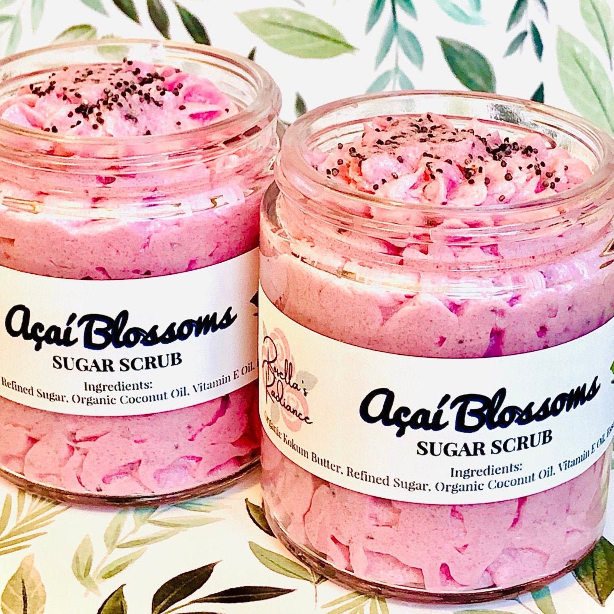 Sugar scrub in glass jars, pink with black specks, "Açaí Blossoms" label visible, on leaf patterned background