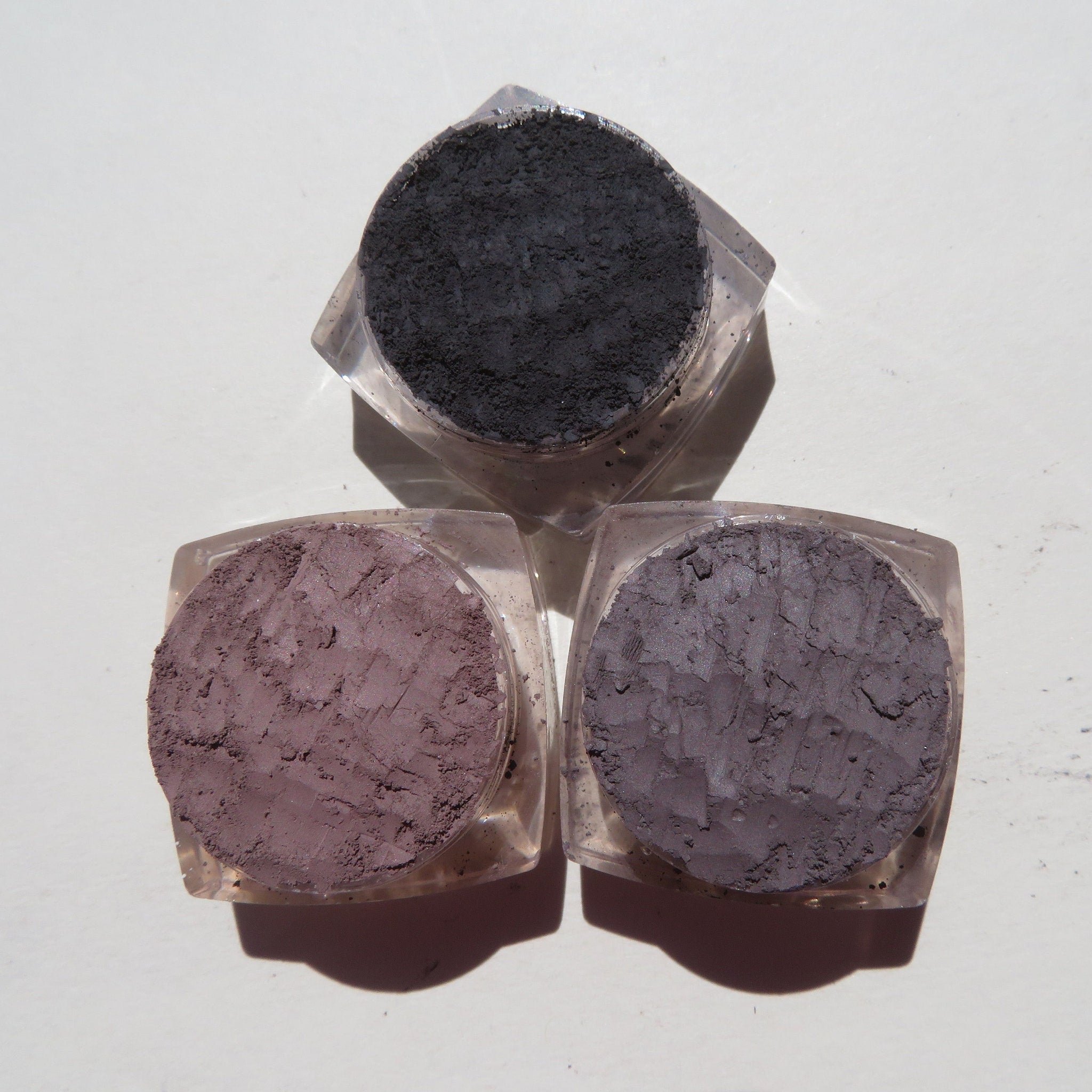 Close-up of four loose mineral eyeshadows in glass jars showing shimmering pink, bronze, taupe, and plum shades