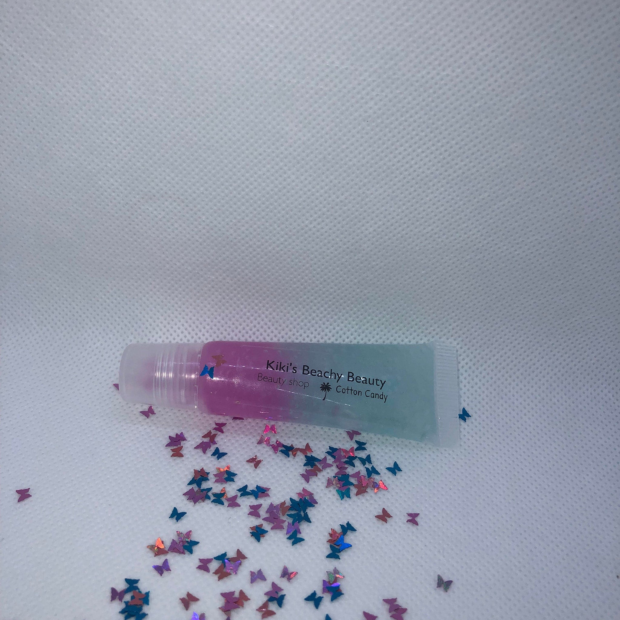 Lip gloss tube with pink and blue gradient, clear tube, and butterfly confetti, Kiki's Beachy Beauty brand