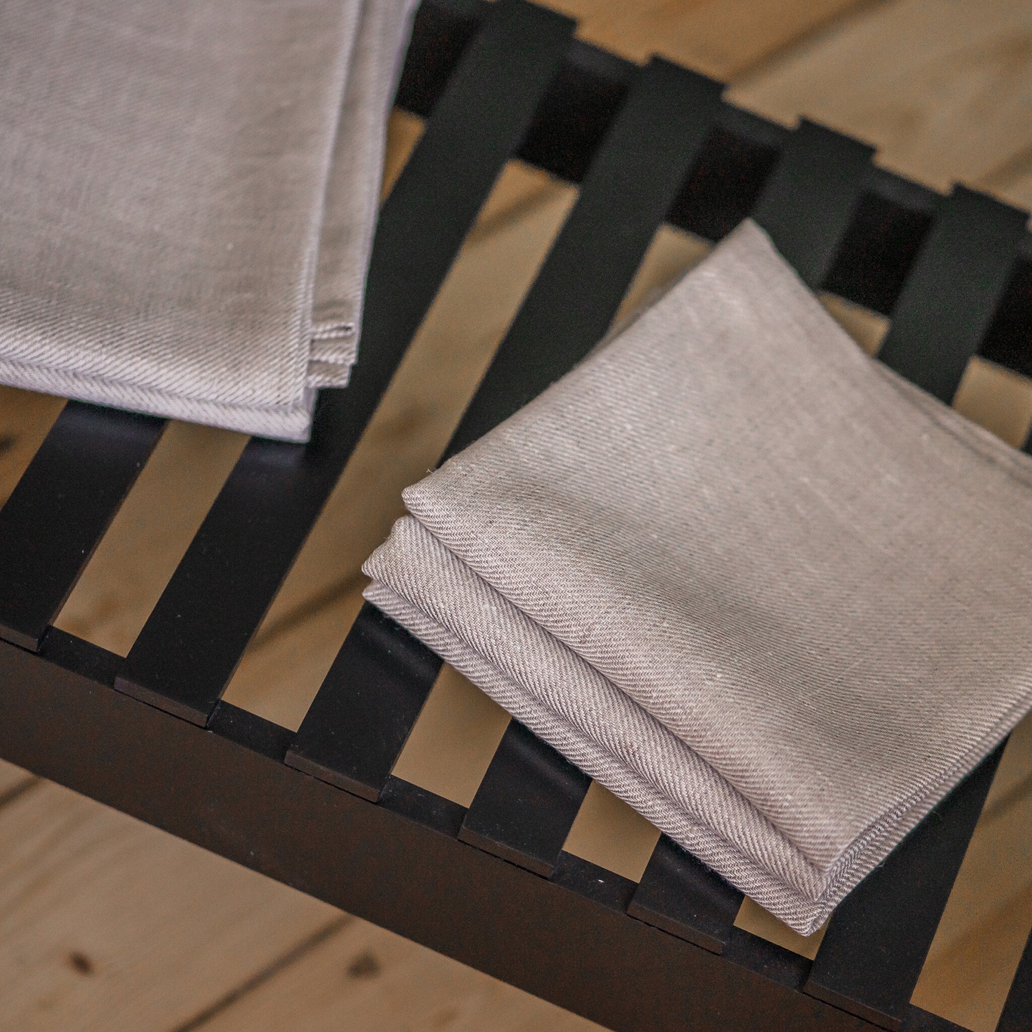 Folded linen napkins, light gray herringbone pattern, stacked on a dark wood slat bed frame