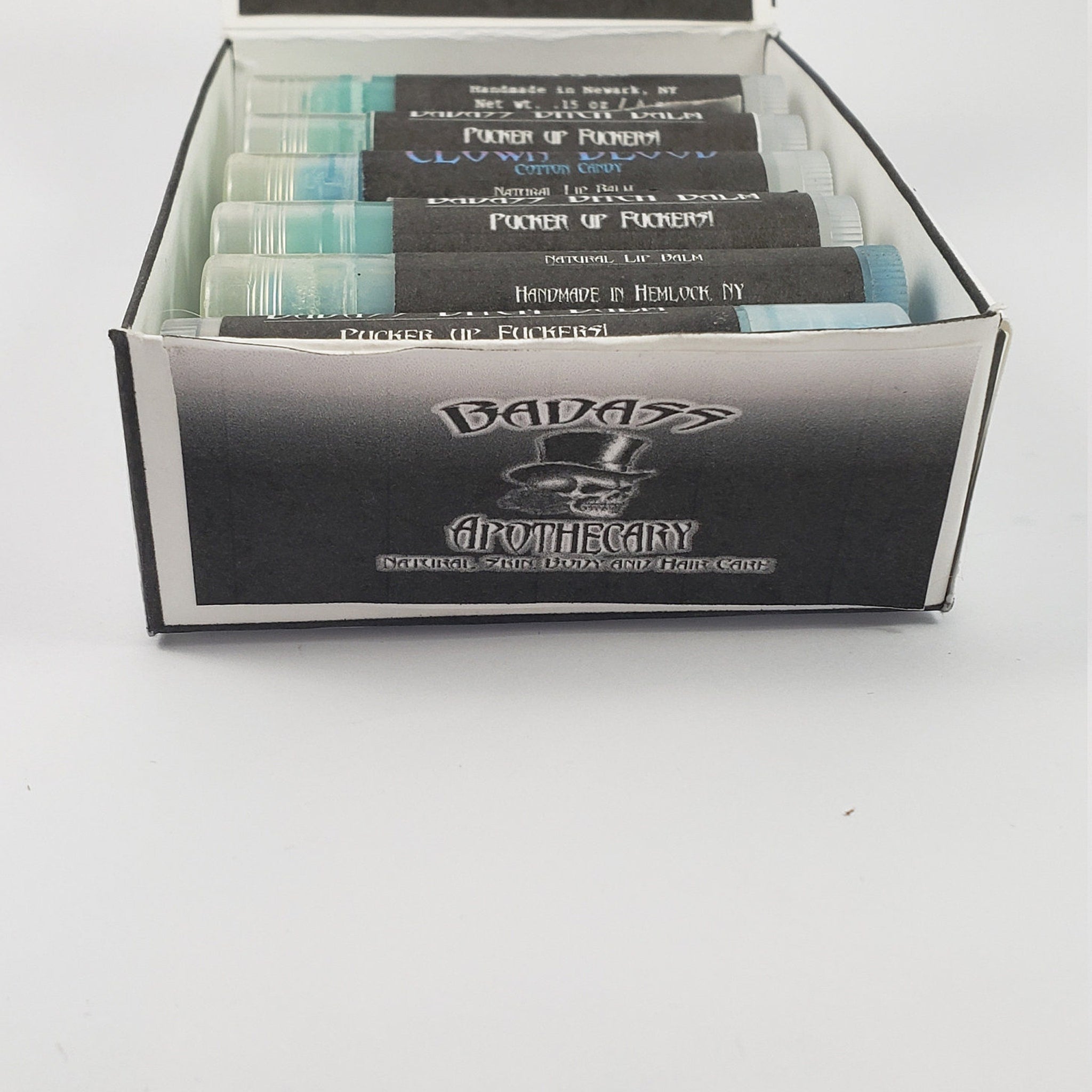 Lip balm tubes with black labels, clear tubes, and BADASS APOTHECARY branding displayed in a box