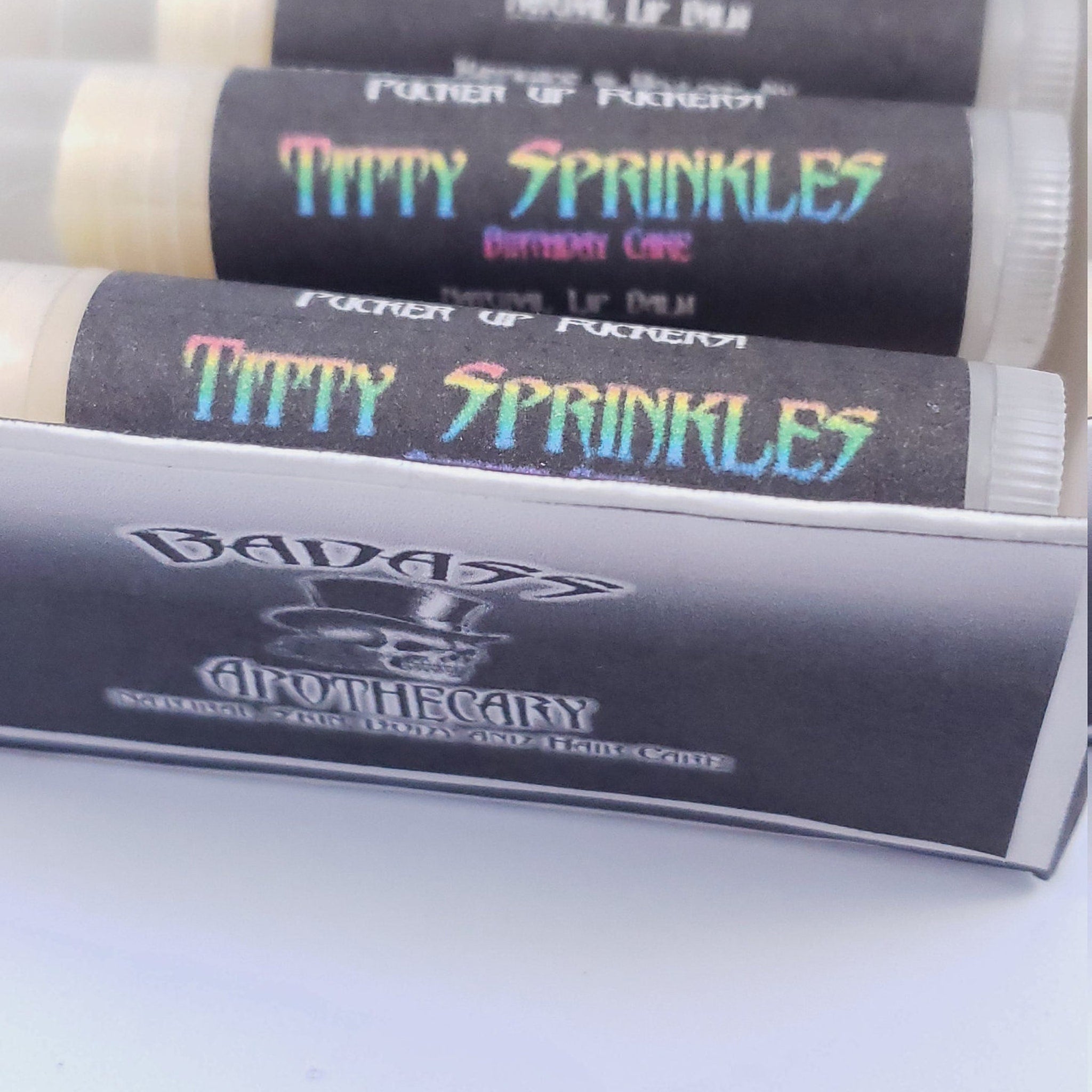 Lip balm tubes with black labels, holographic text, and white containers, "Titty Sprinkles" brand name visible