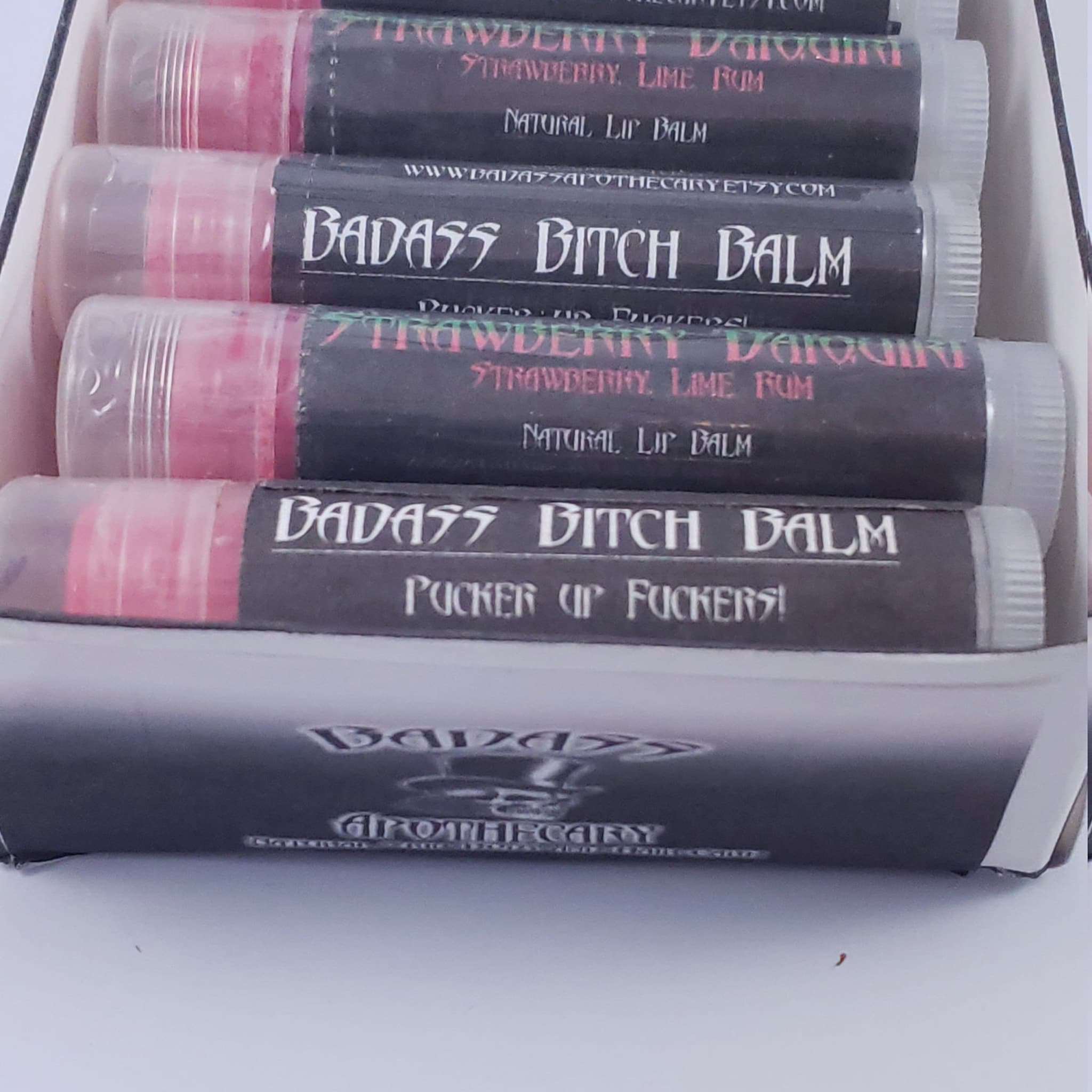 Lip balm tubes with pink balm, black labels, and clear caps, "BADASS BITCH BALM" text visible