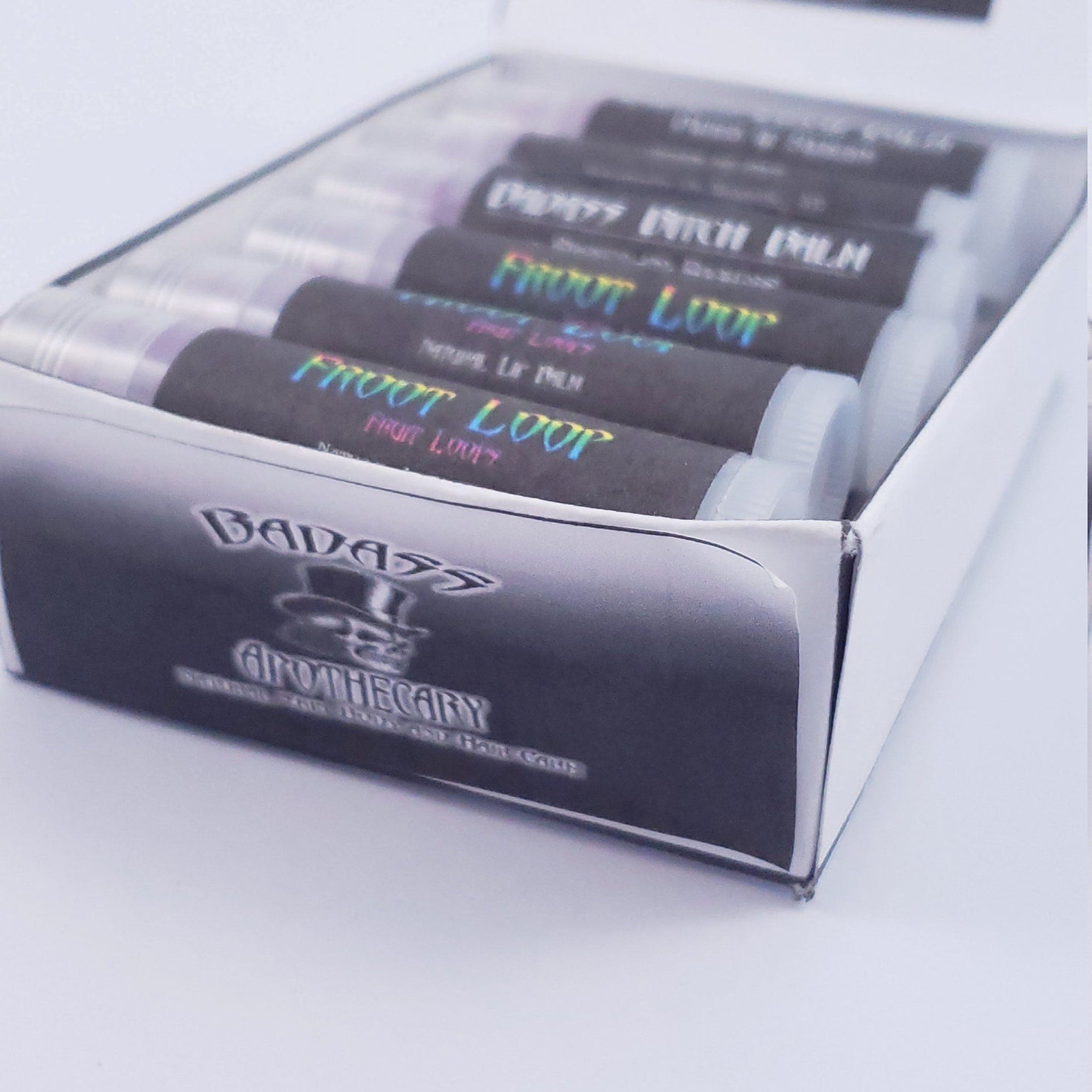 Lip balm tubes with black labels, white caps, and holographic text, in a white box, Badass Apothecary brand