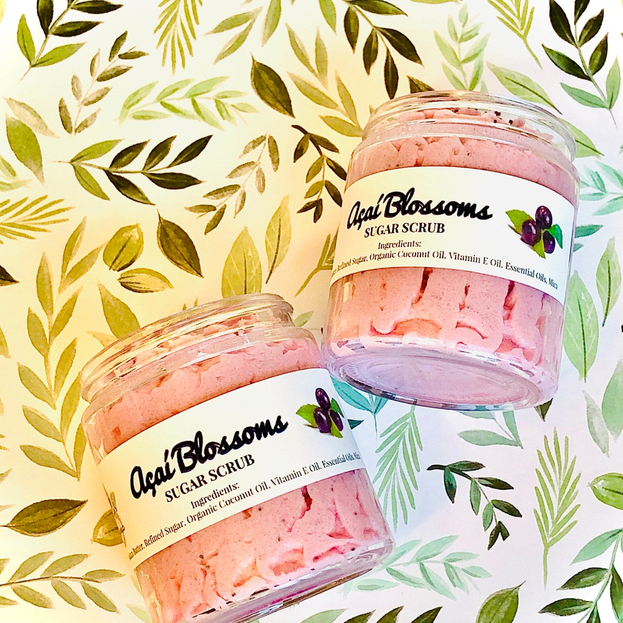 Sugar scrub jars with pink scrub, clear glass, and "Açaí Blossoms" label on green leaf background.
