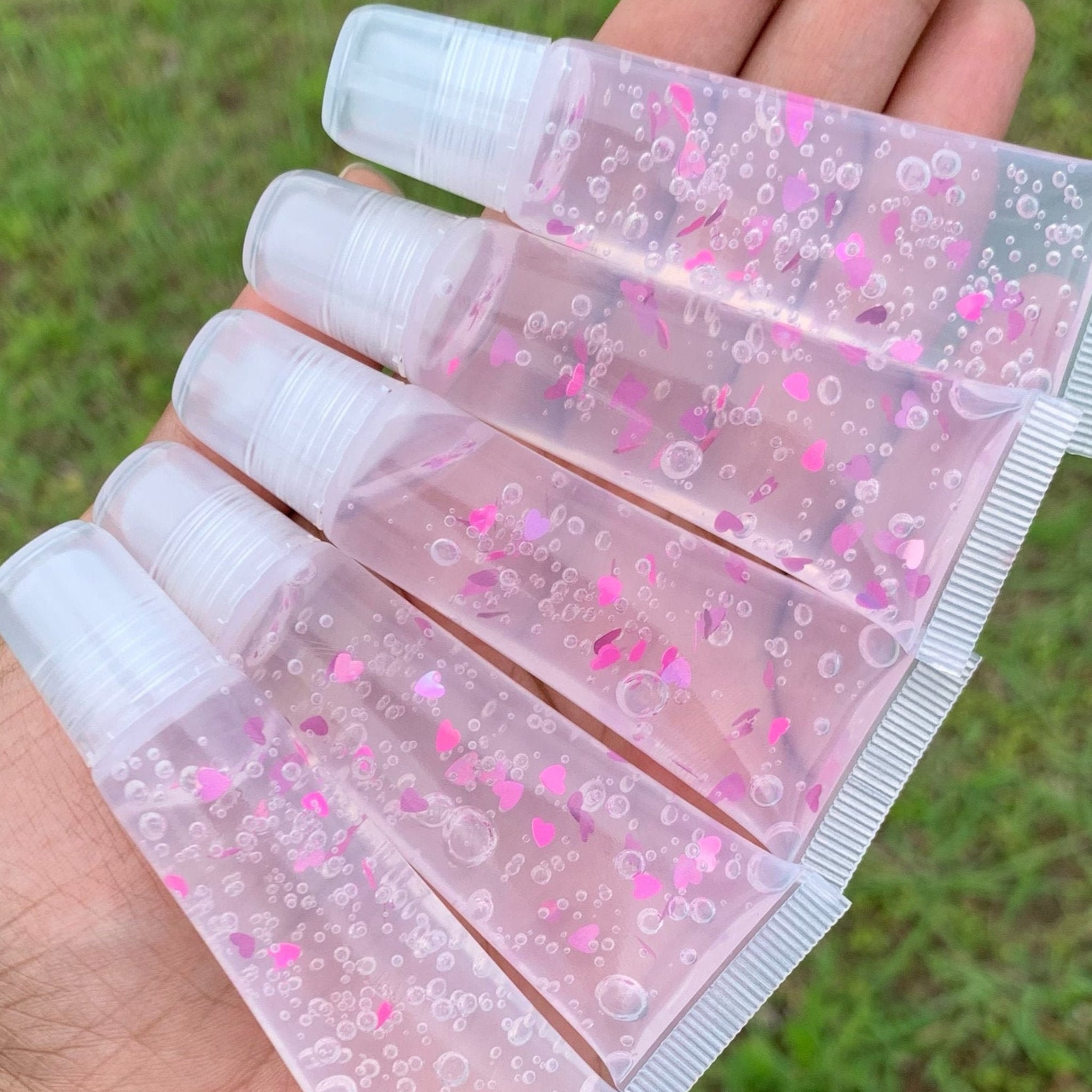 Lip gloss tubes with clear liquid, pink heart-shaped glitter, and clear caps held in hand