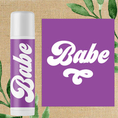 Lip balm tube with purple label, white text "Babe", and white cap, on burlap background
