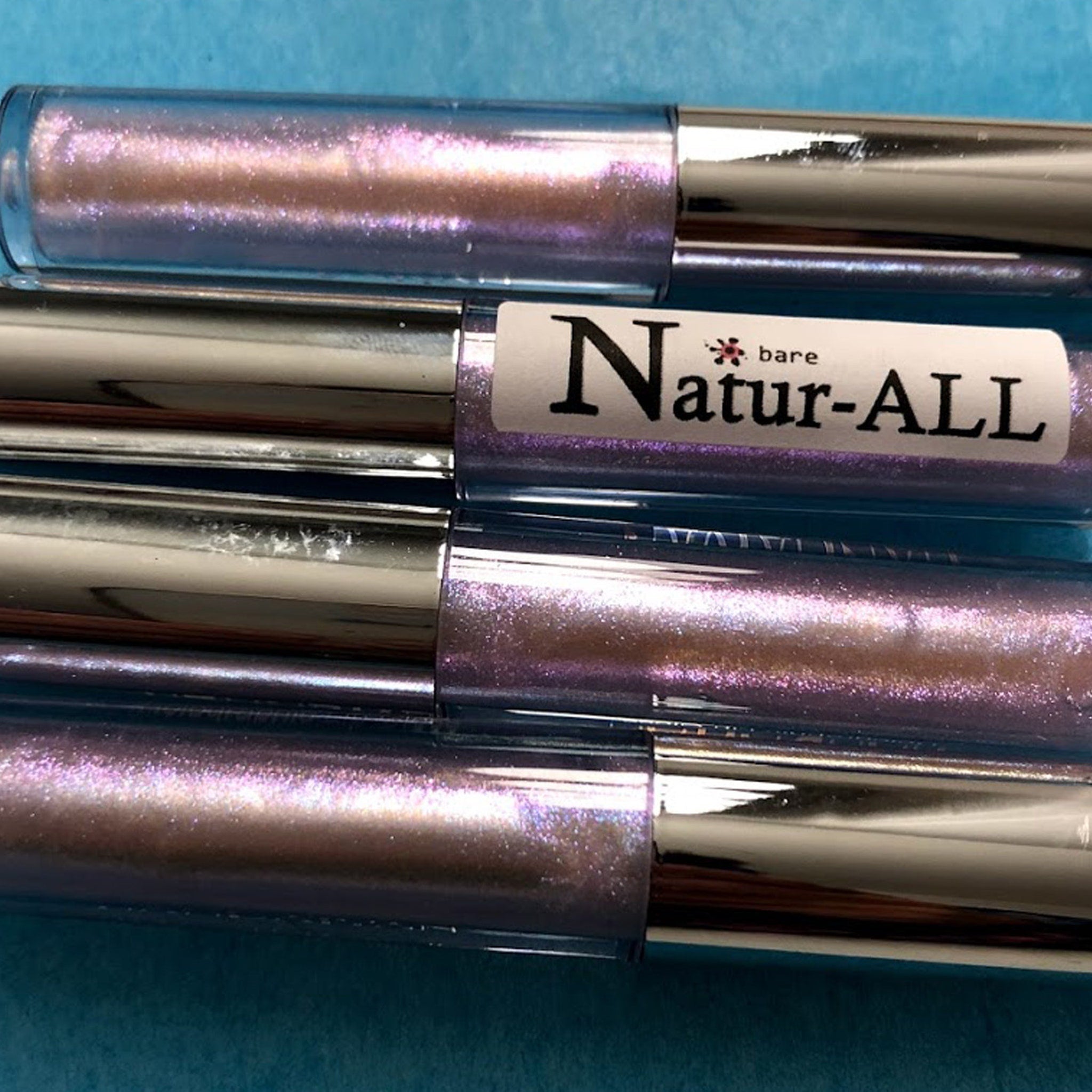 Lip gloss tubes with iridescent purple, silver cap, and "Natur-ALL" label on blue surface