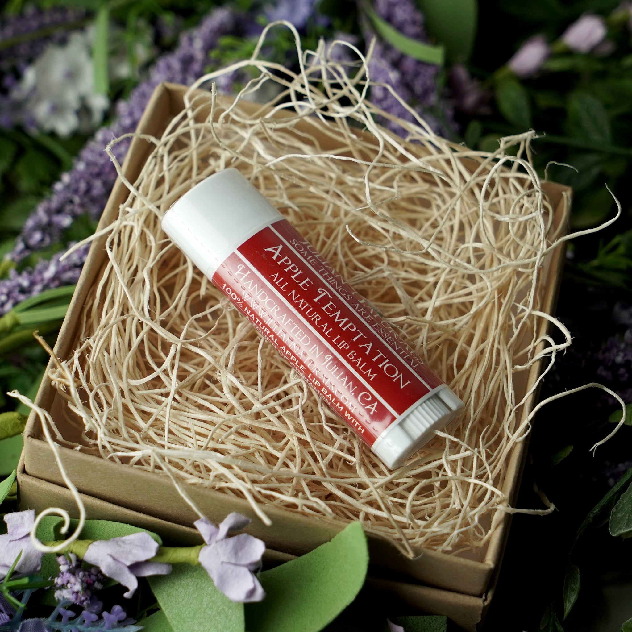 Lip balm tube with red and white label, in a box with wood shavings, Apple Temptation brand