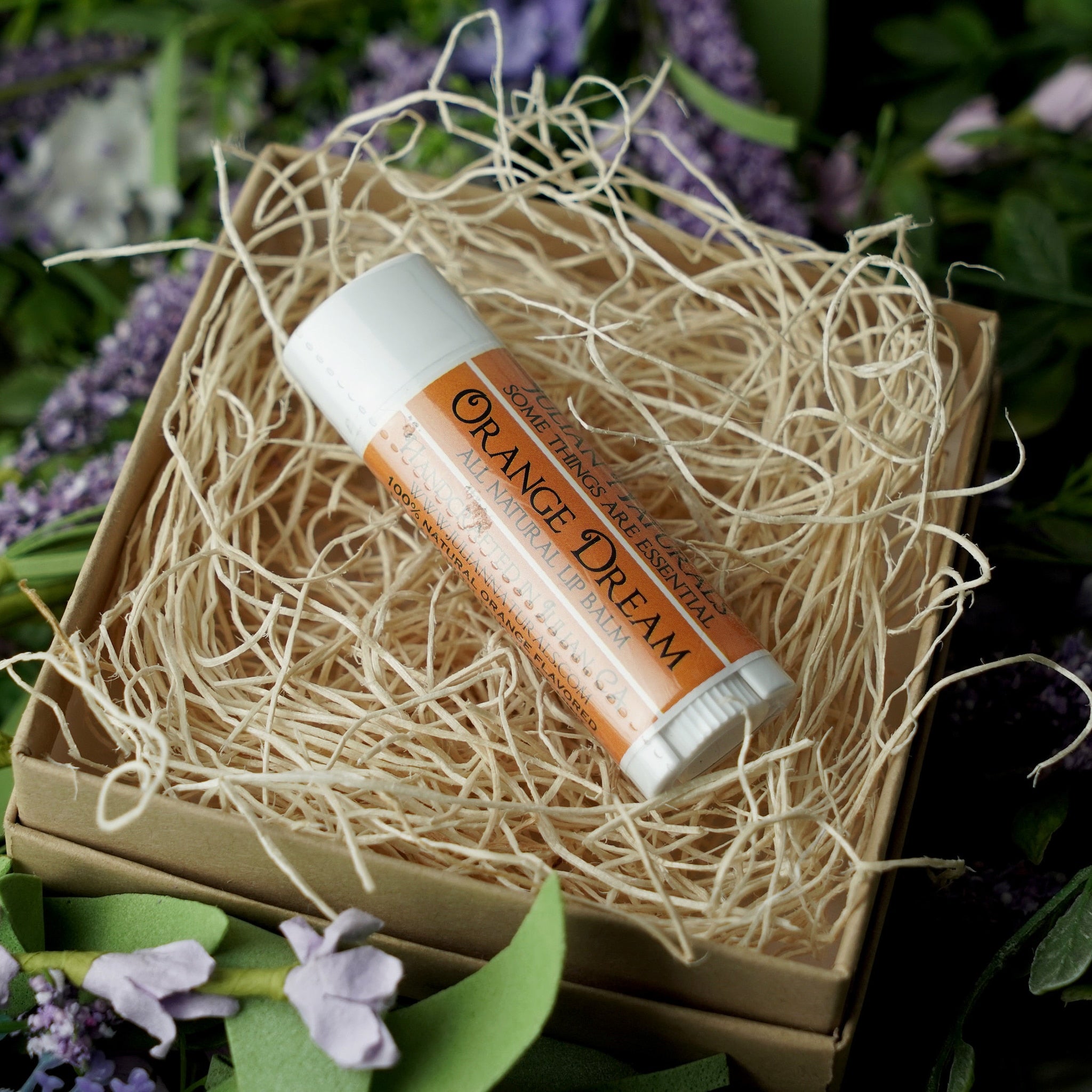 Orange Dream lip balm tube with white and orange label, in a gift box with straw filler, surrounded by flowers