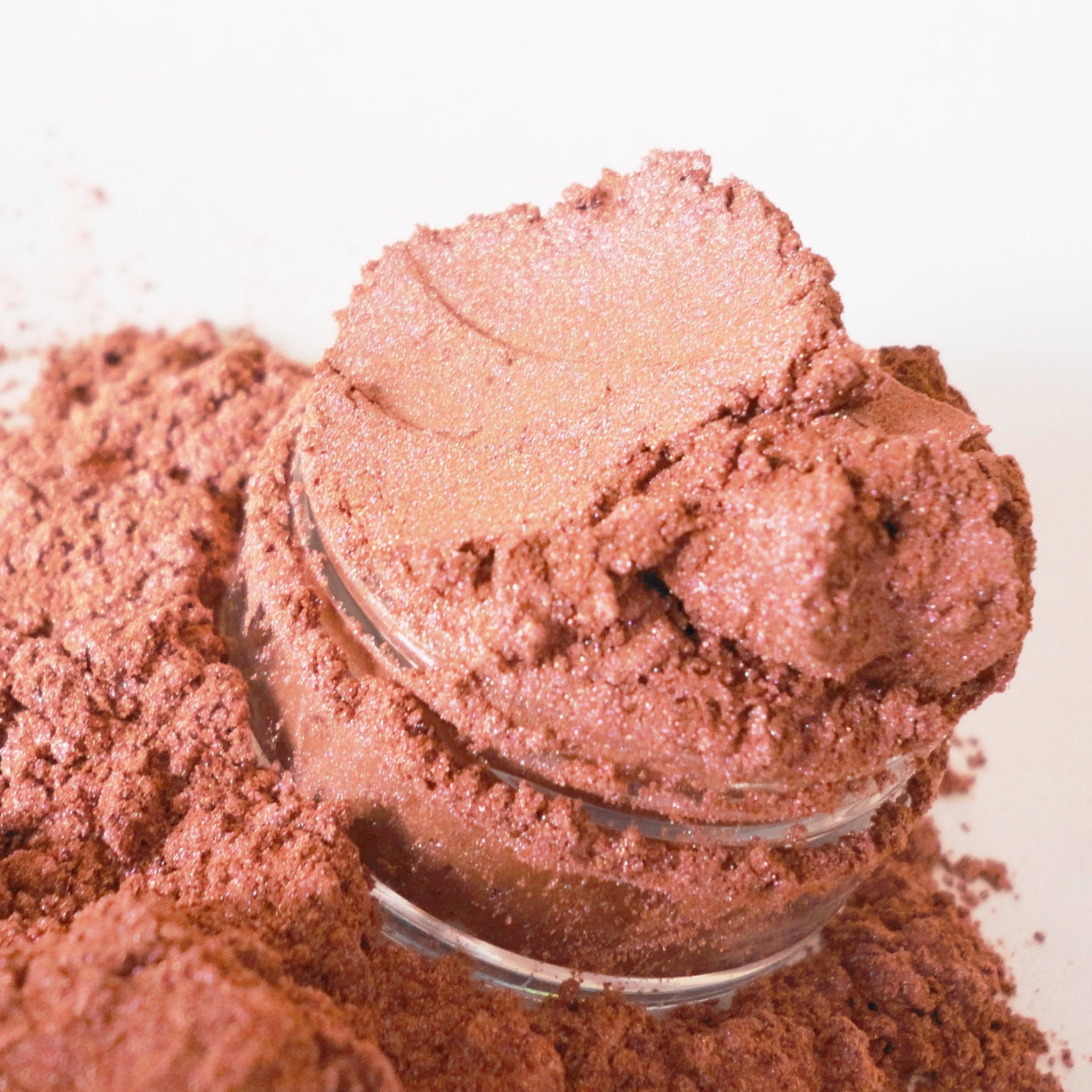 Powdered eyeshadow in rose-gold color with shimmer, in a glass container and spilled around it.