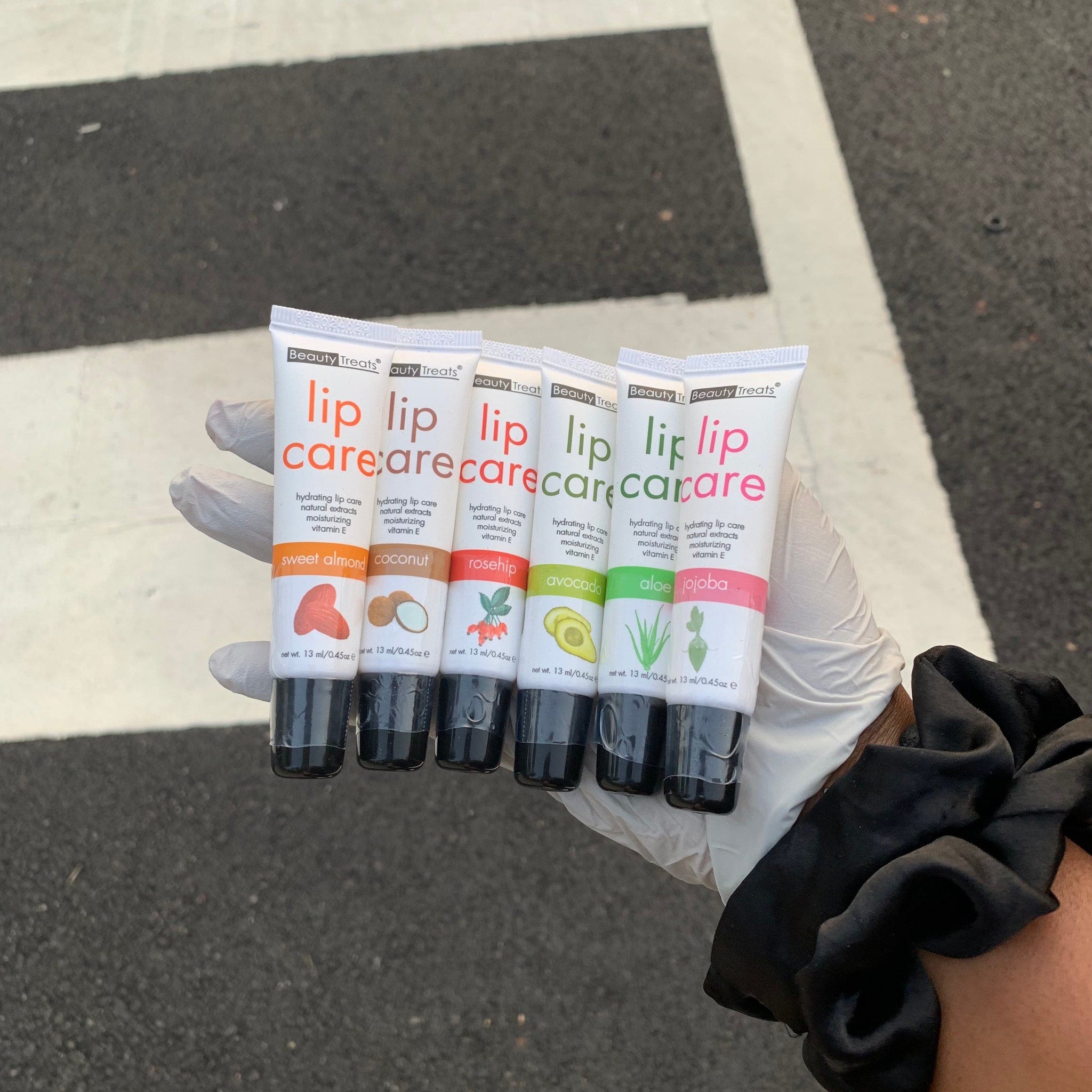Lip care tubes in white and black, various flavors, held in gloved hand, Beauty Treats brand