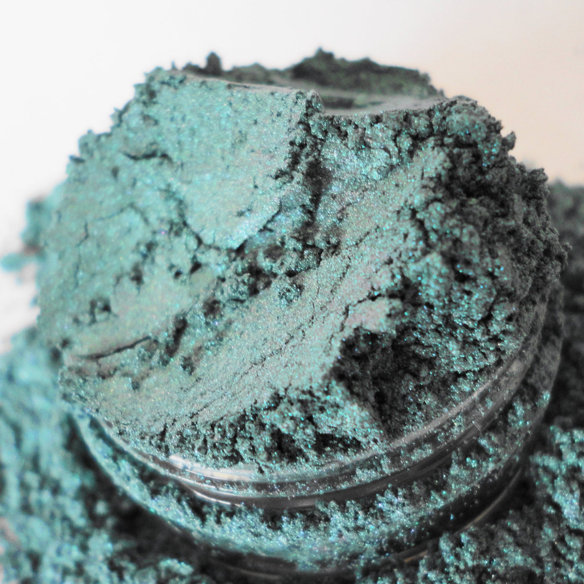 Eyeshadow powder in teal color with glittery texture and container visible