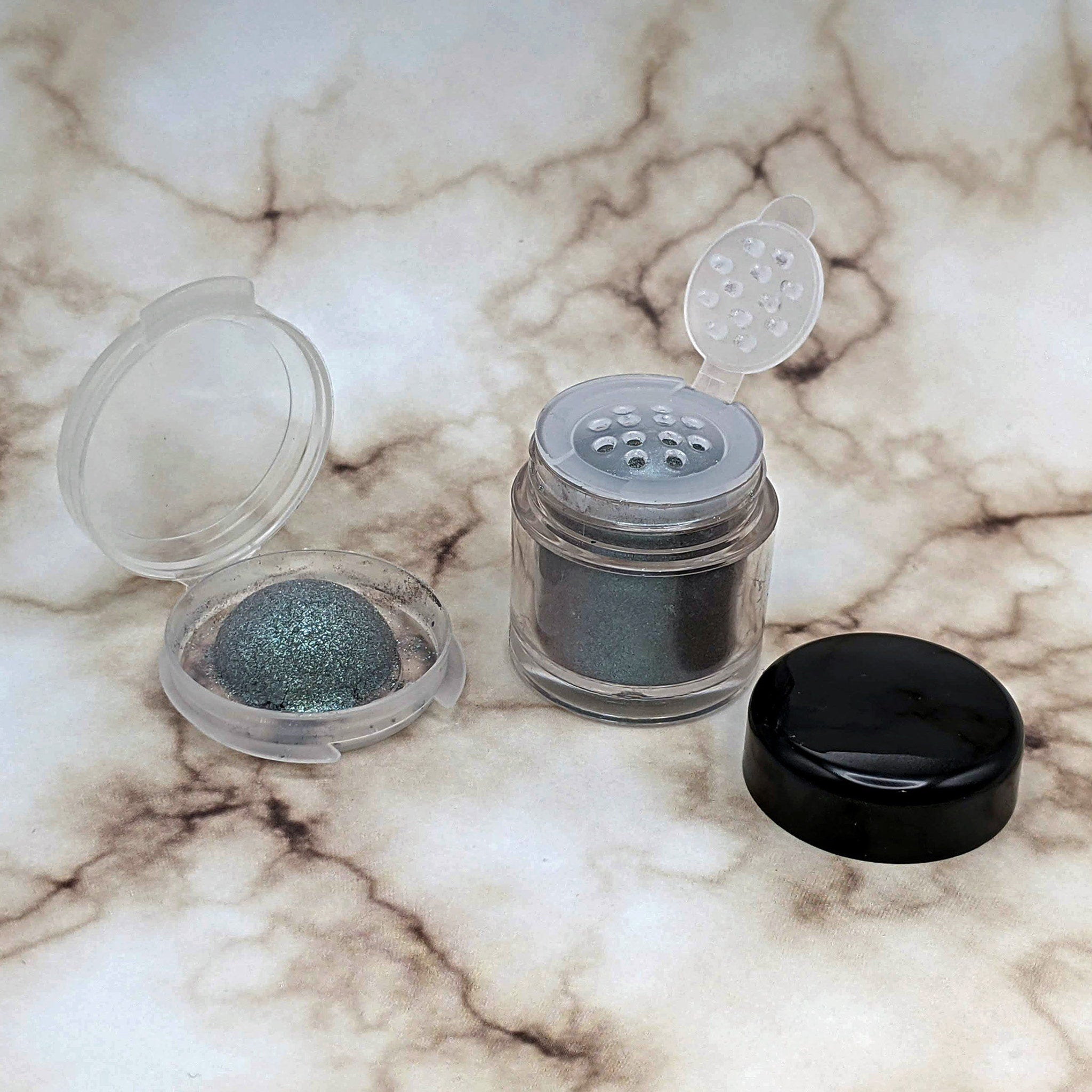 Eyeshadow powder in clear and black containers with green shimmer and marble background