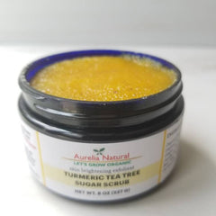 Close-up of golden turmeric scrub with visible tea tree leaves in a textured glass jar, showcasing natural exfoliants