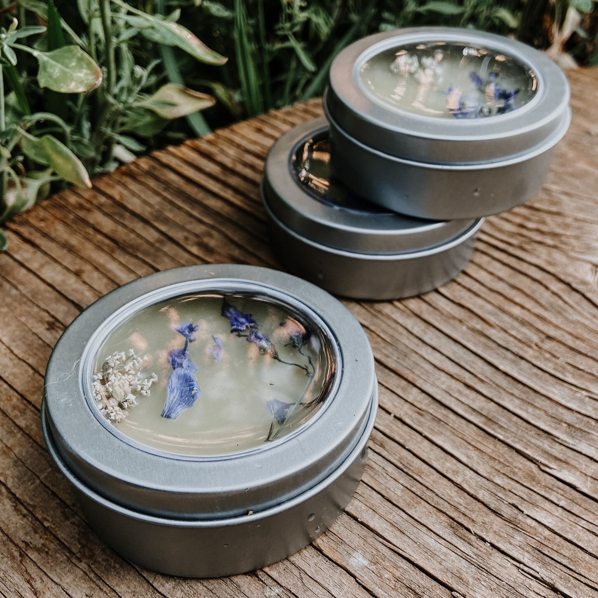 Candle tins with clear lids, silver metal, and floral accents on wooden surface