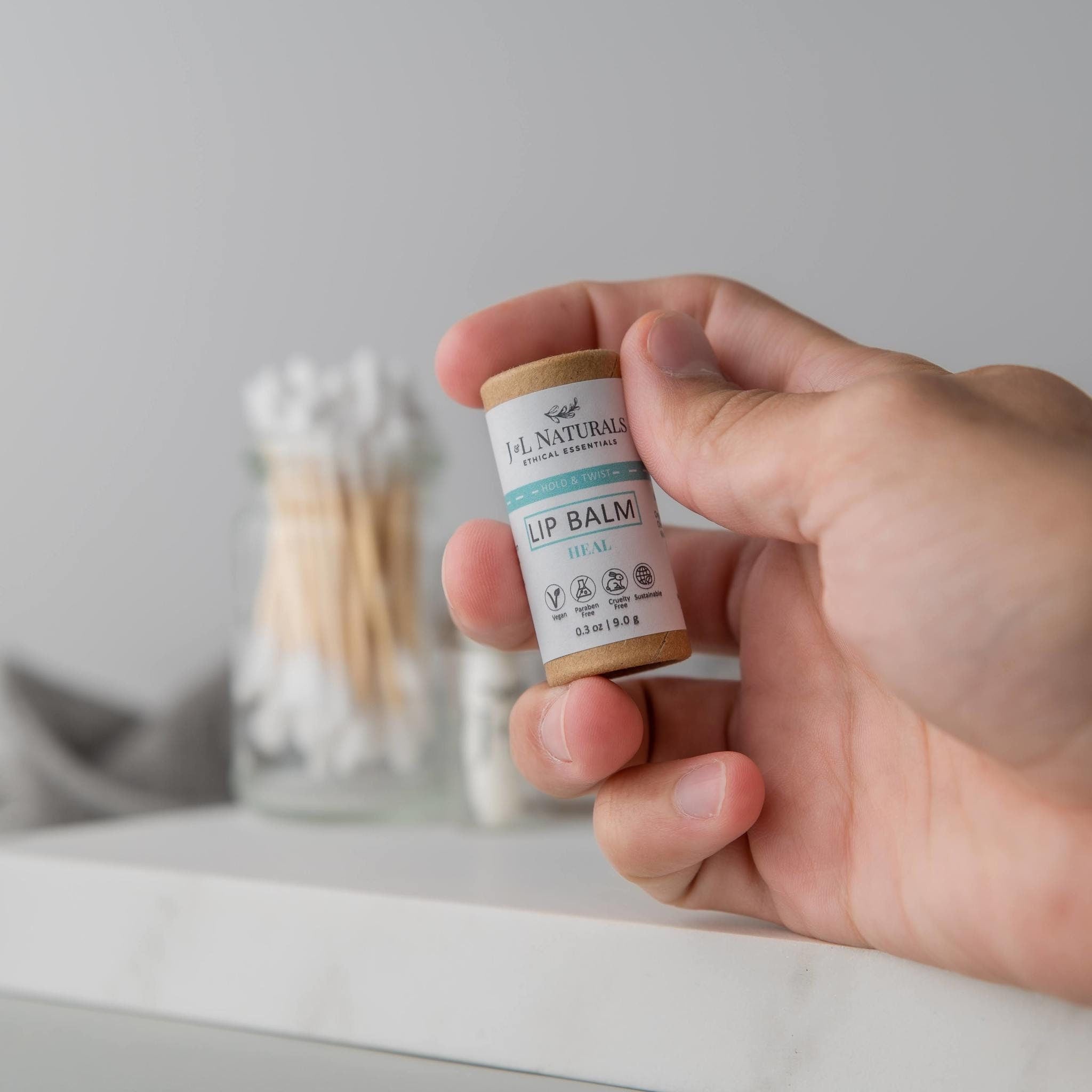 Lip balm tube in hand, brown cardboard with white label, J&L Naturals branding visible