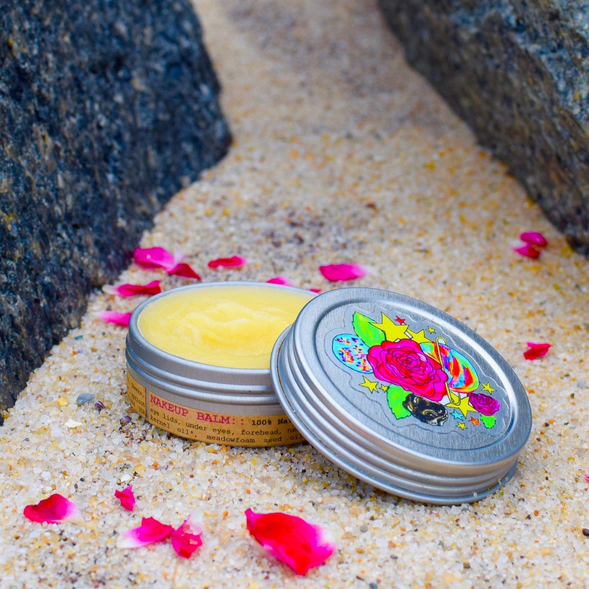 Lip balm in silver tin with floral lid design and yellow balm, on sand with rose petals