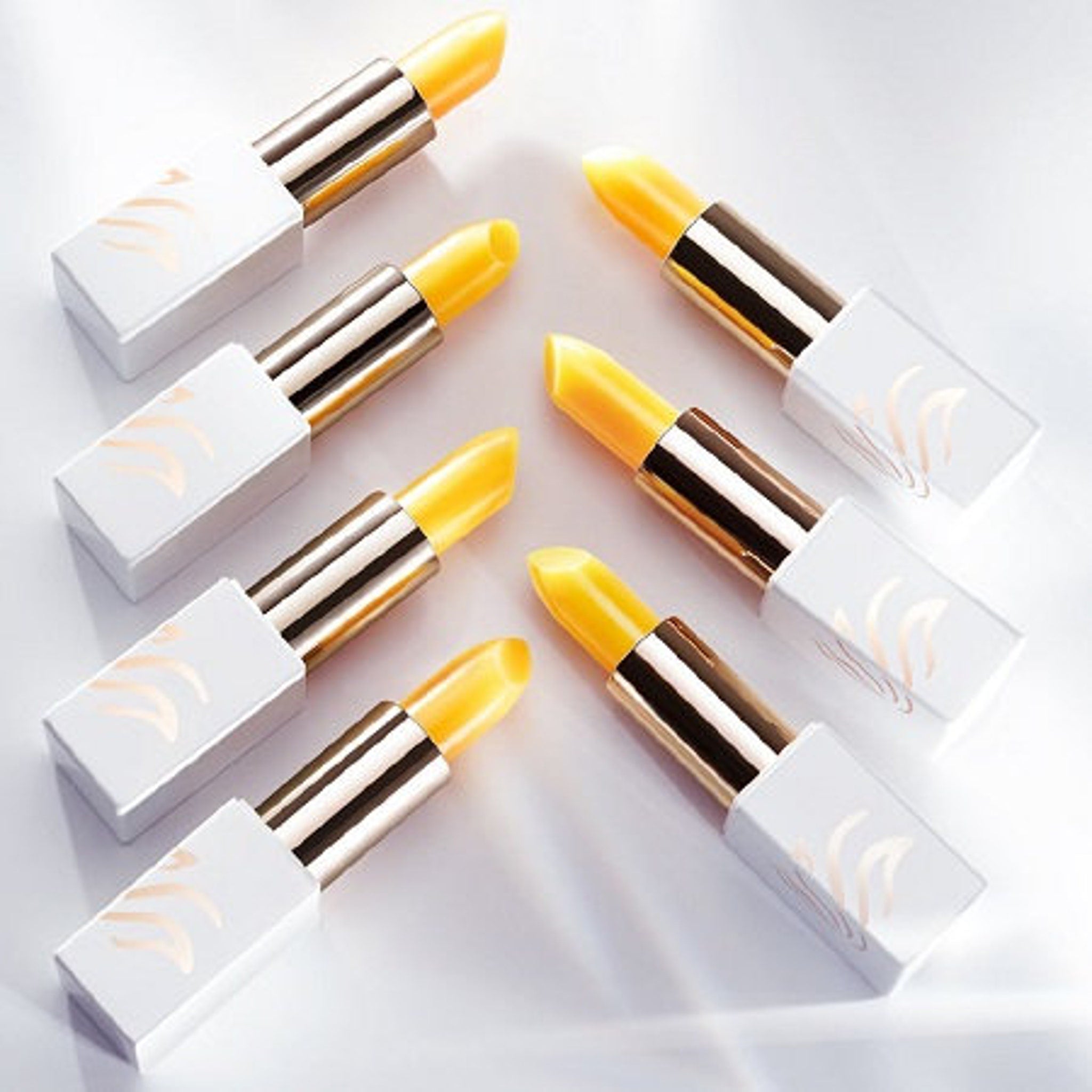 Lipsticks with yellow balm, white and gold casing, and gold logo detail, arranged on white surface