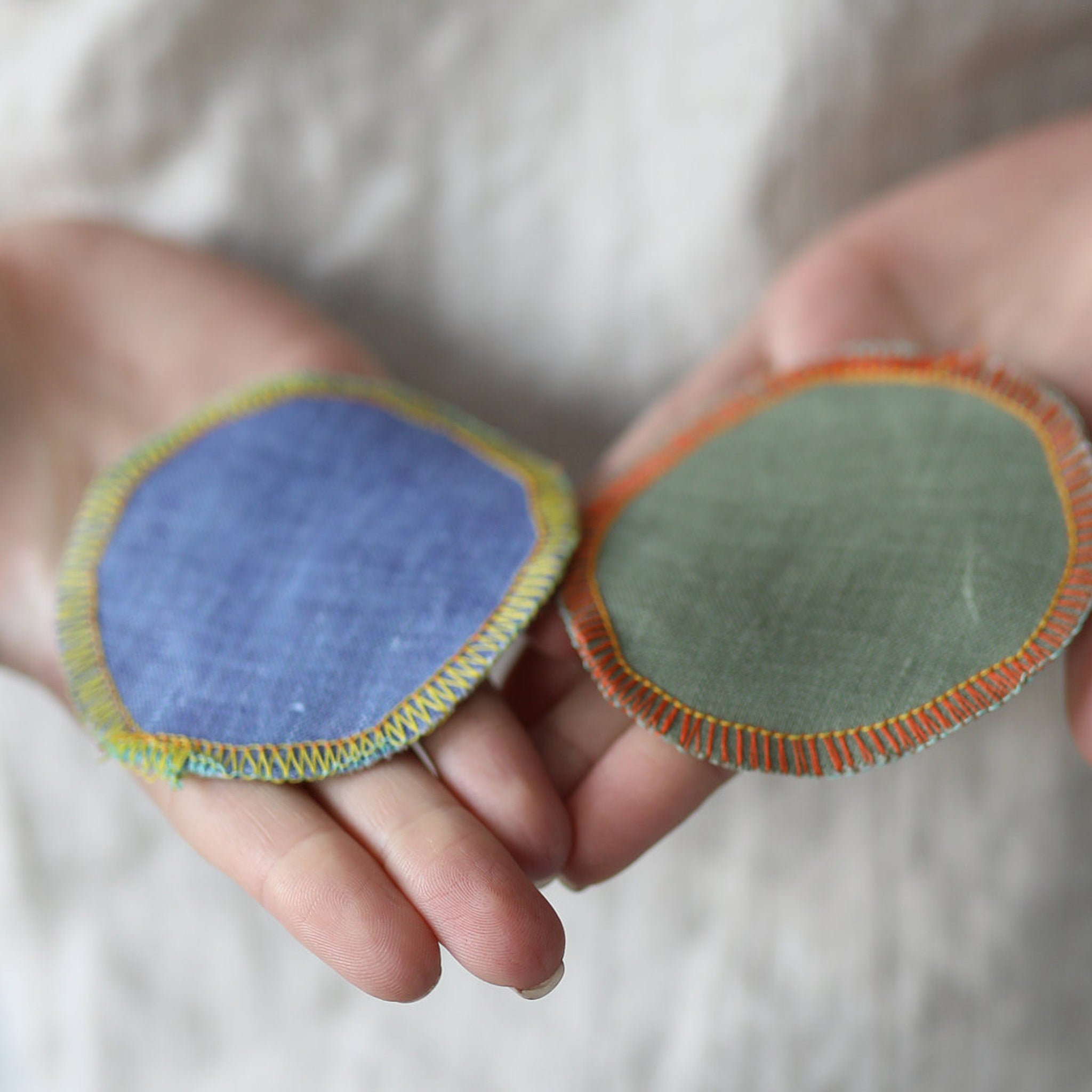 Reusable makeup remover pads, blue and green linen rounds with colorful stitching, held in hands