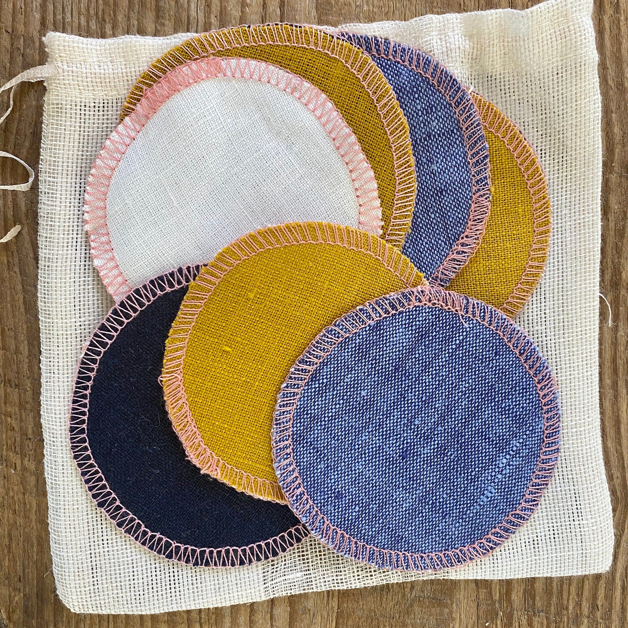 Reusable facial rounds in various colors including blue, yellow, and white with pink stitching, in a mesh bag