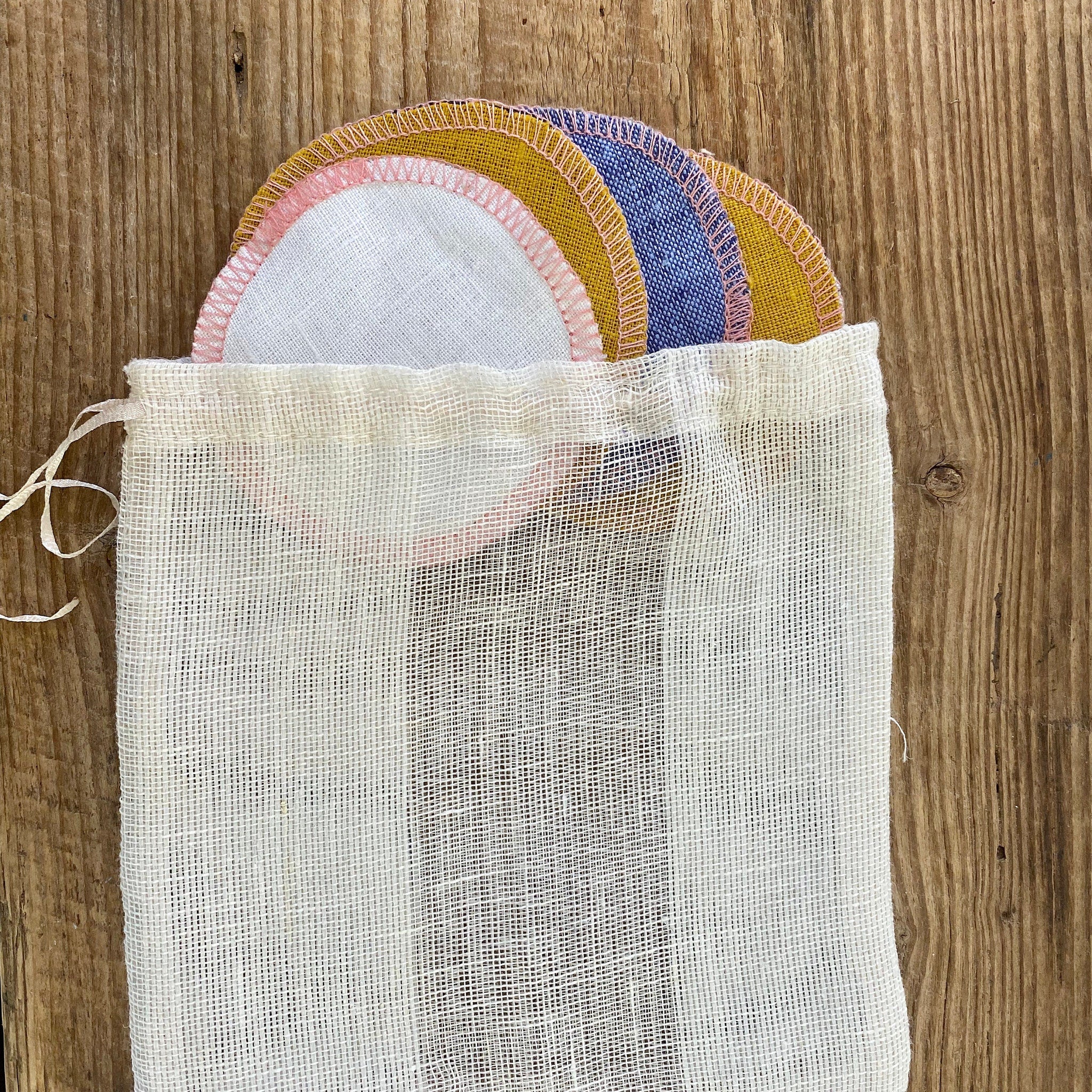 Reusable facial rounds in various colors, white linen bag with drawstring, wooden background