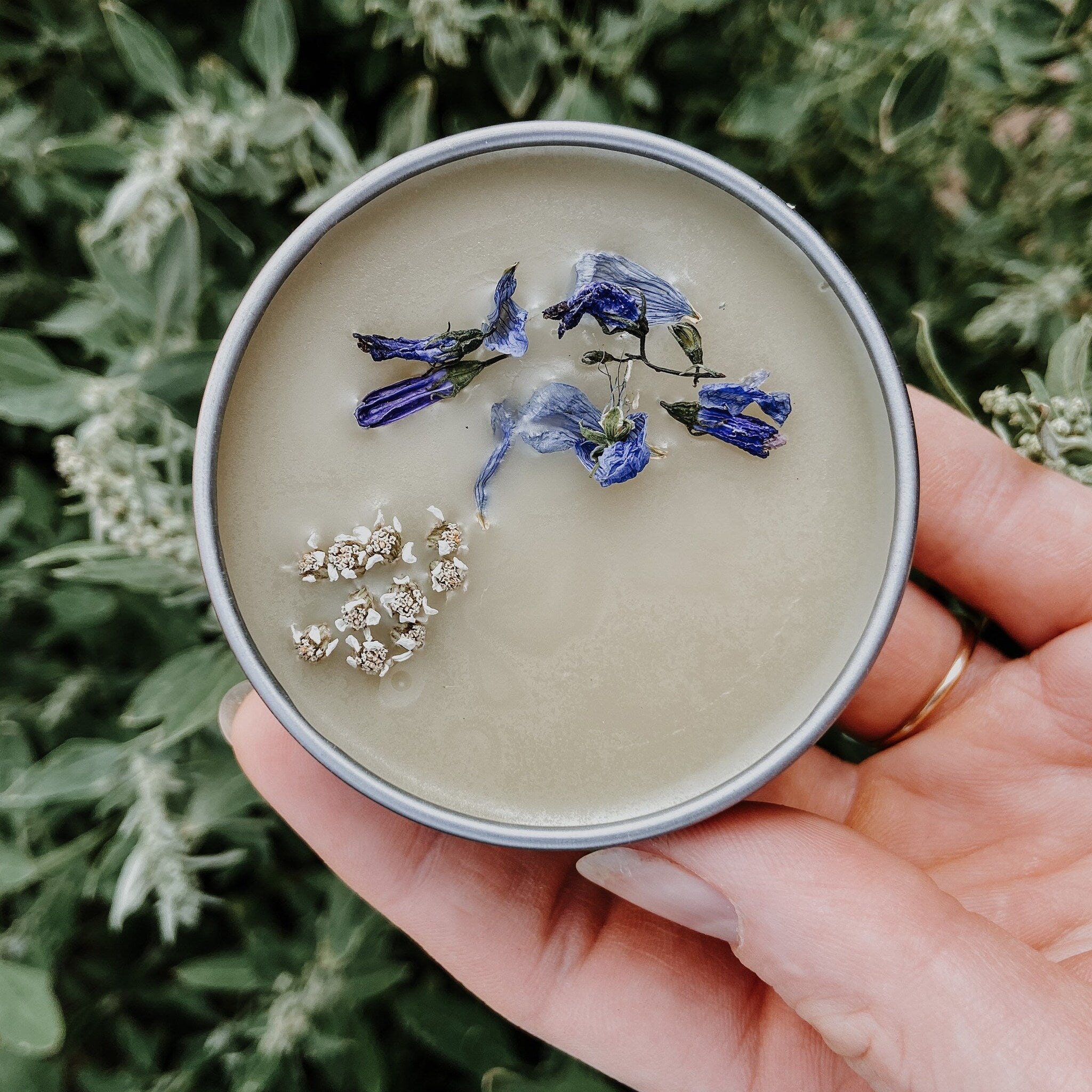 Cream-colored balm with blue and white flowers in a silver tin held in a hand