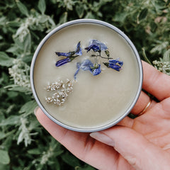 Cream-colored balm with blue and white flowers in a silver tin held in a hand