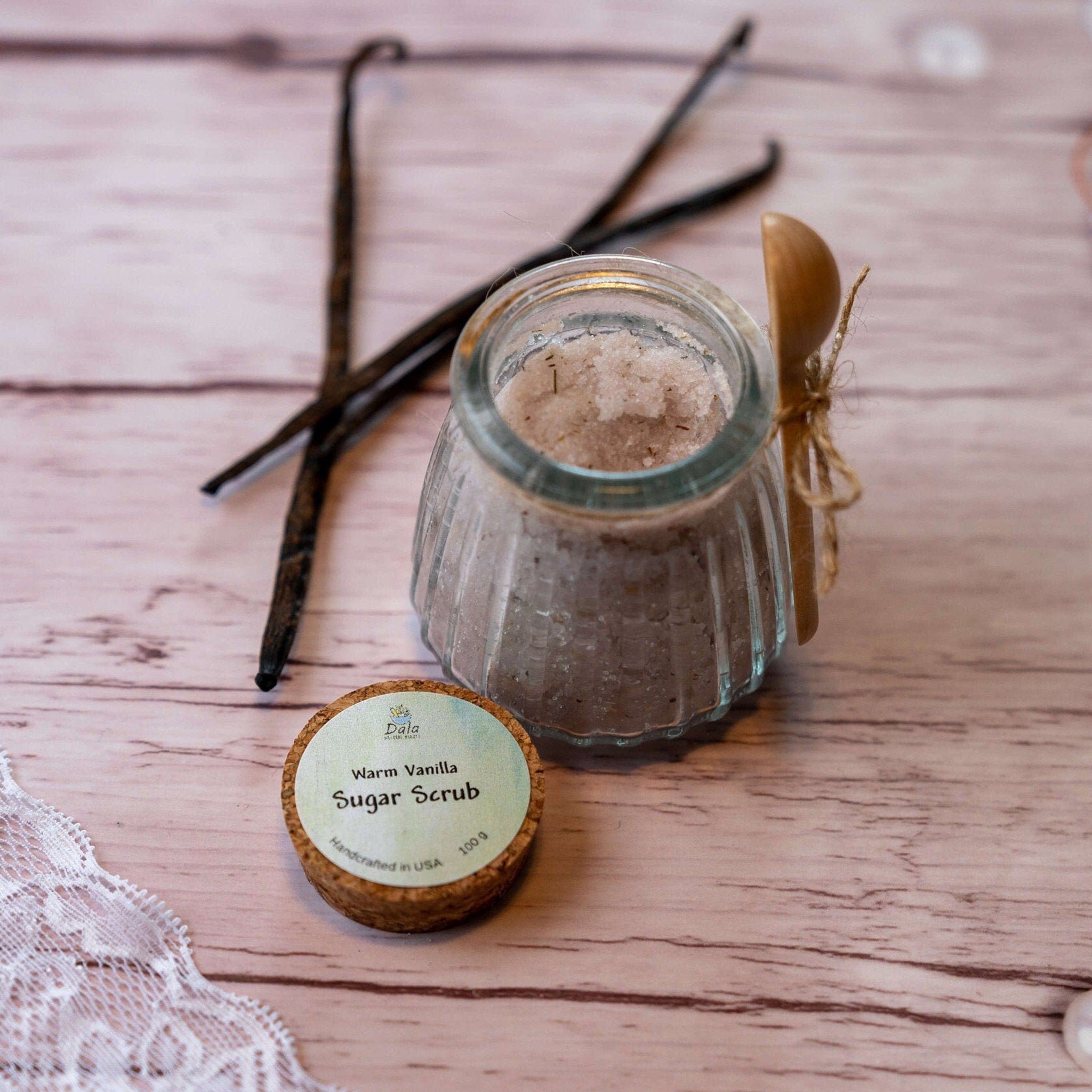 Sugar scrub in glass jar with wooden spoon and vanilla beans on wood surface, DalaNaturalBeauty label