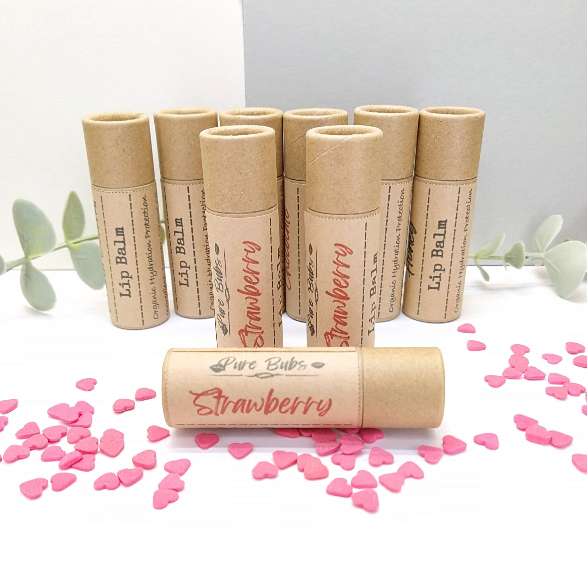 Lip balm tubes in brown cardboard with strawberry label and pink heart sprinkles on white surface
